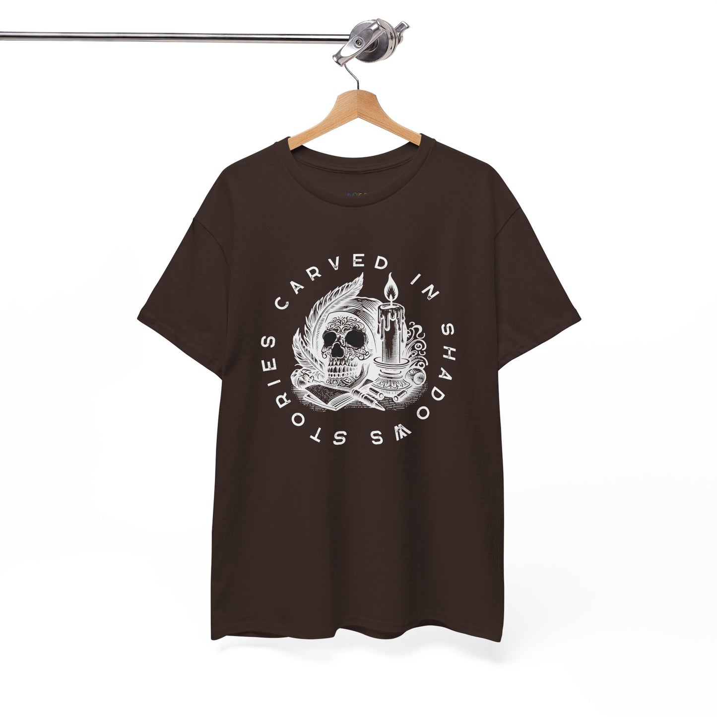 Brown t-shirt with white graphic design of a creepy skull, books, candle and feathers with text 'Stories Carved In Shadows' on a hanger against a white background