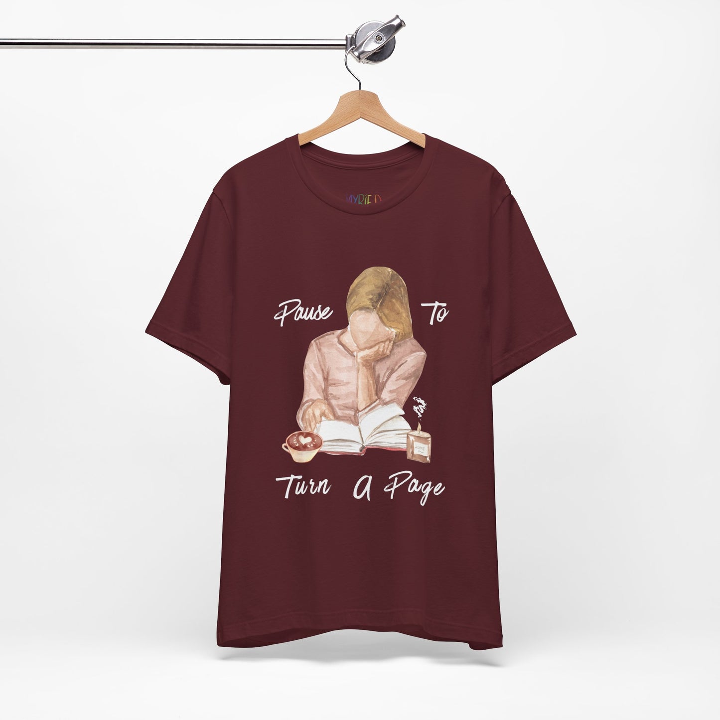 Maroon t-shirt with a graphic design of a woman reading a book, a hot beverage, candle and text 'Pause to Turn a Page'