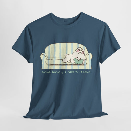 Blue t-shirt with a white graphic bunny, pink ears, reading a green book on a blue and yellow striped couch, text below 'some bunny loves to learn'