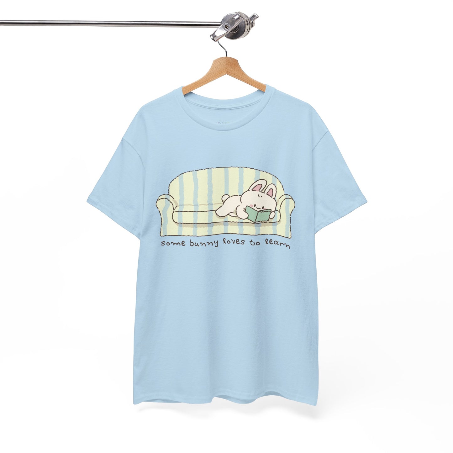 Light blue t-shirt with a cartoon bunny reading a book as it's lays on a couch, text 'some bunny loves to learn'