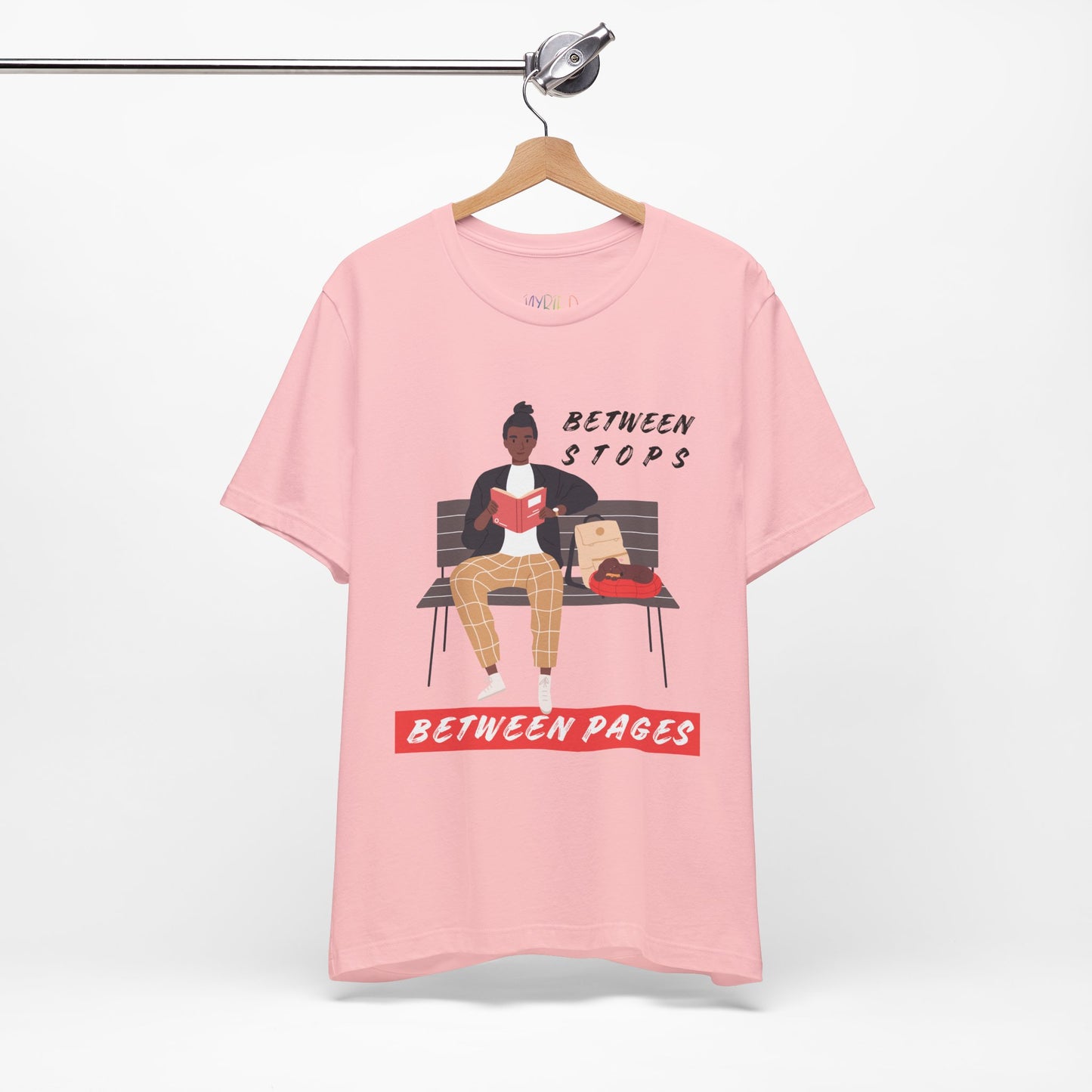 Pink t-shirt with a graphic of a person sitting on a bench reading a book, with text 'Between Stops Between Pages'