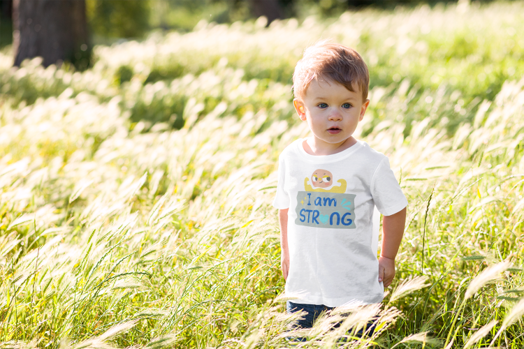 Toddler in a grassy field wearing a white t-shirt with 'I Am Strong' text by Jaybie D.