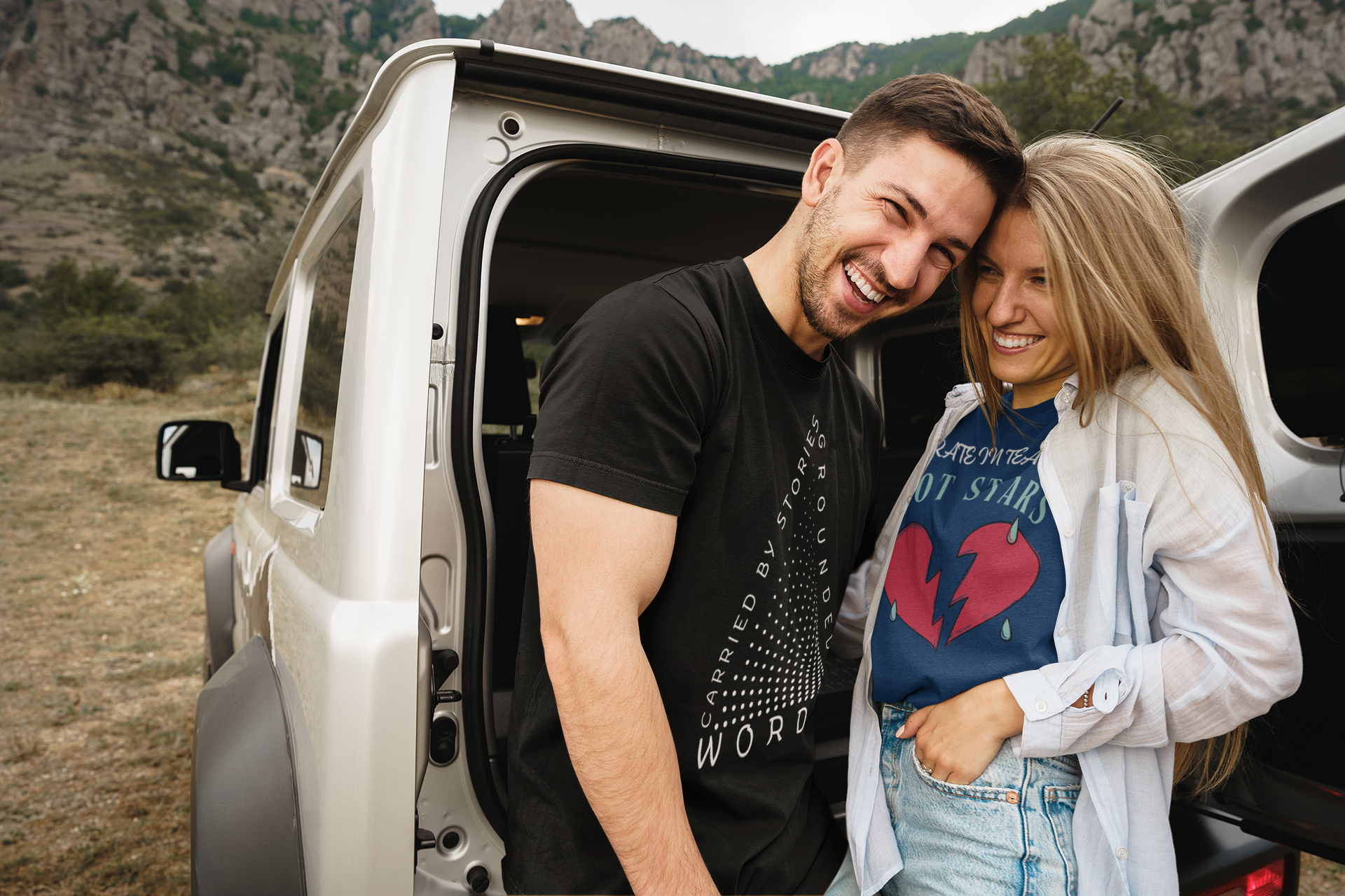 A man and a woman on a nature adventure, standing behind their vehicle with an open door, and wearing t-shirts by Jaybie D.