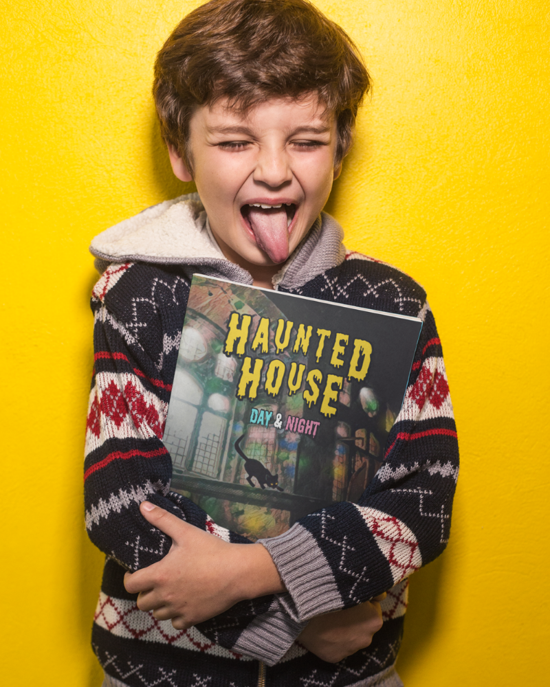 Child sticking out his tongue while holding a book titled 'Haunted House Day & Night' by Jaybie D. as he stands against a yellow background