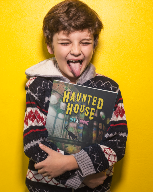Young boy making a silly face with his tongue sticking out, as he stands against a yellow wall holding picture book Haunted House Day & Night by Jaybie D.