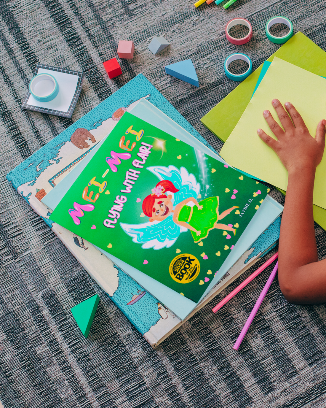 Child laying on the ground with crafts and a copy of children's picture book Mei-Mei Flying With Flair by Jaybie D.