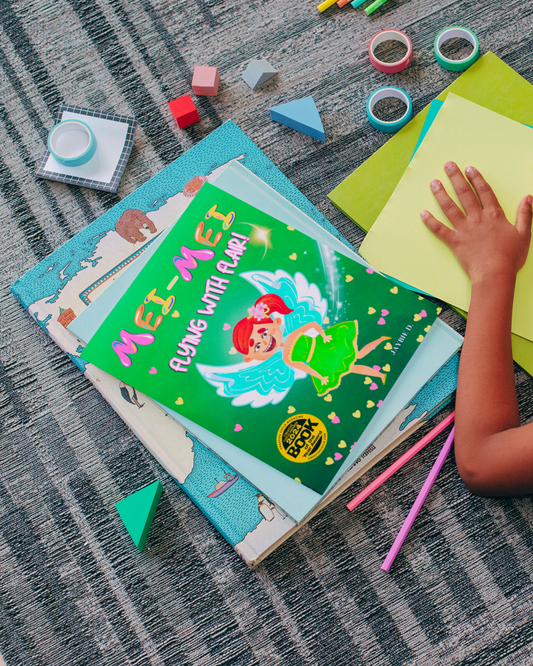 Child laying on the ground with crafts and a copy of children's picture book Mei-Mei Flying With Flair by Jaybie D.