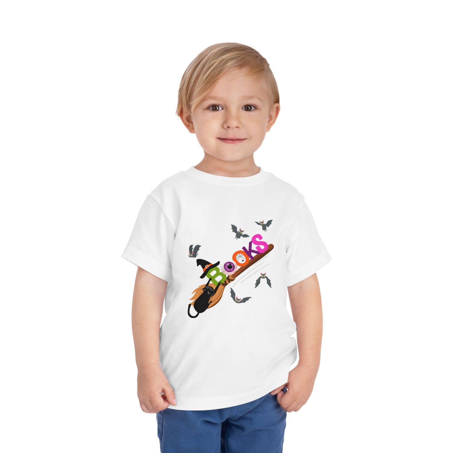 Child wearing a white toddler t-shirt with colorful graphic design of 'BOOKS' riding a broom with a black cat barely handing on and bats all around