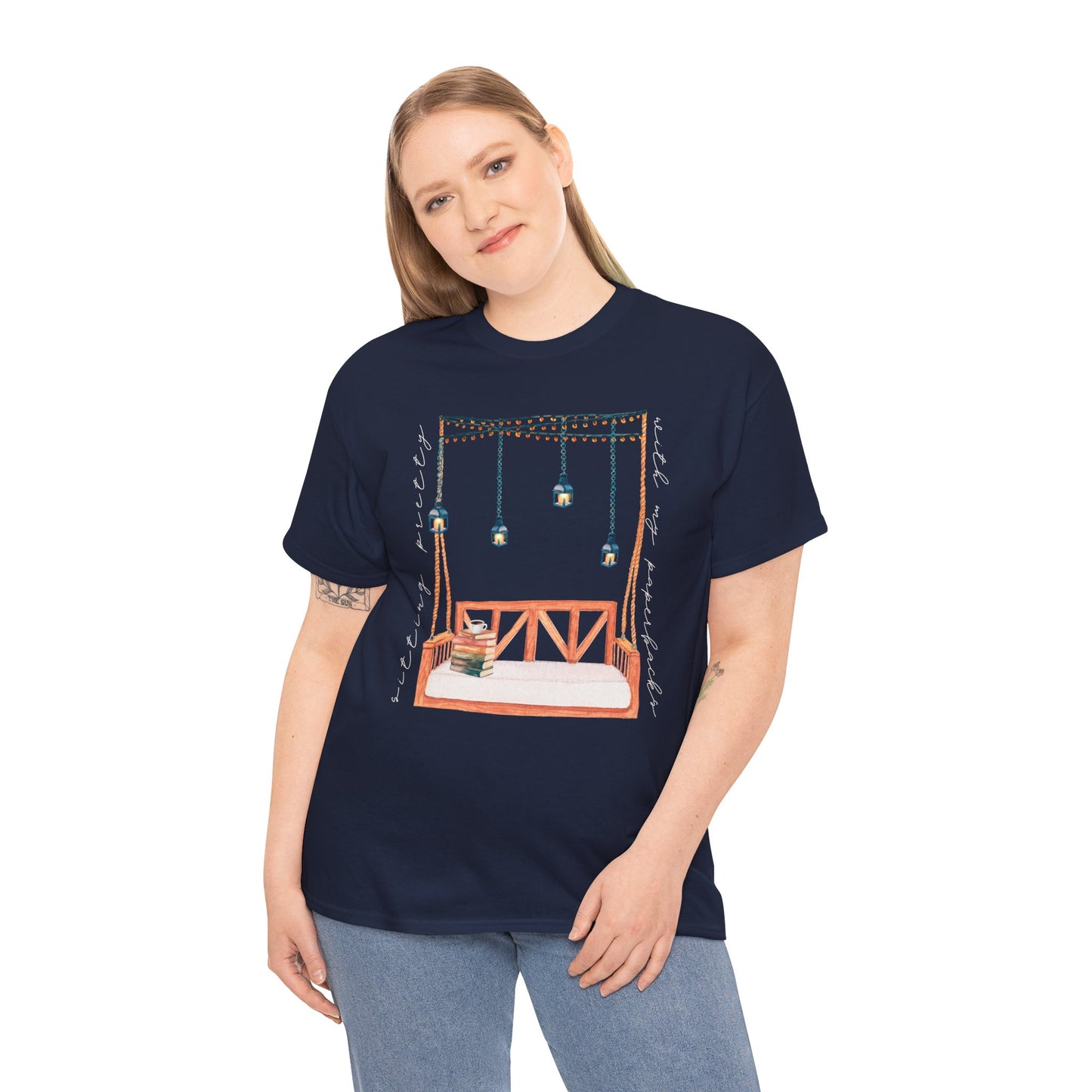 Woman wearing a navy blue t-shirt with a porch swing, string lights, hot beverage and a stack of books, text says 'Sitting Pretty With My Paperbacks'
