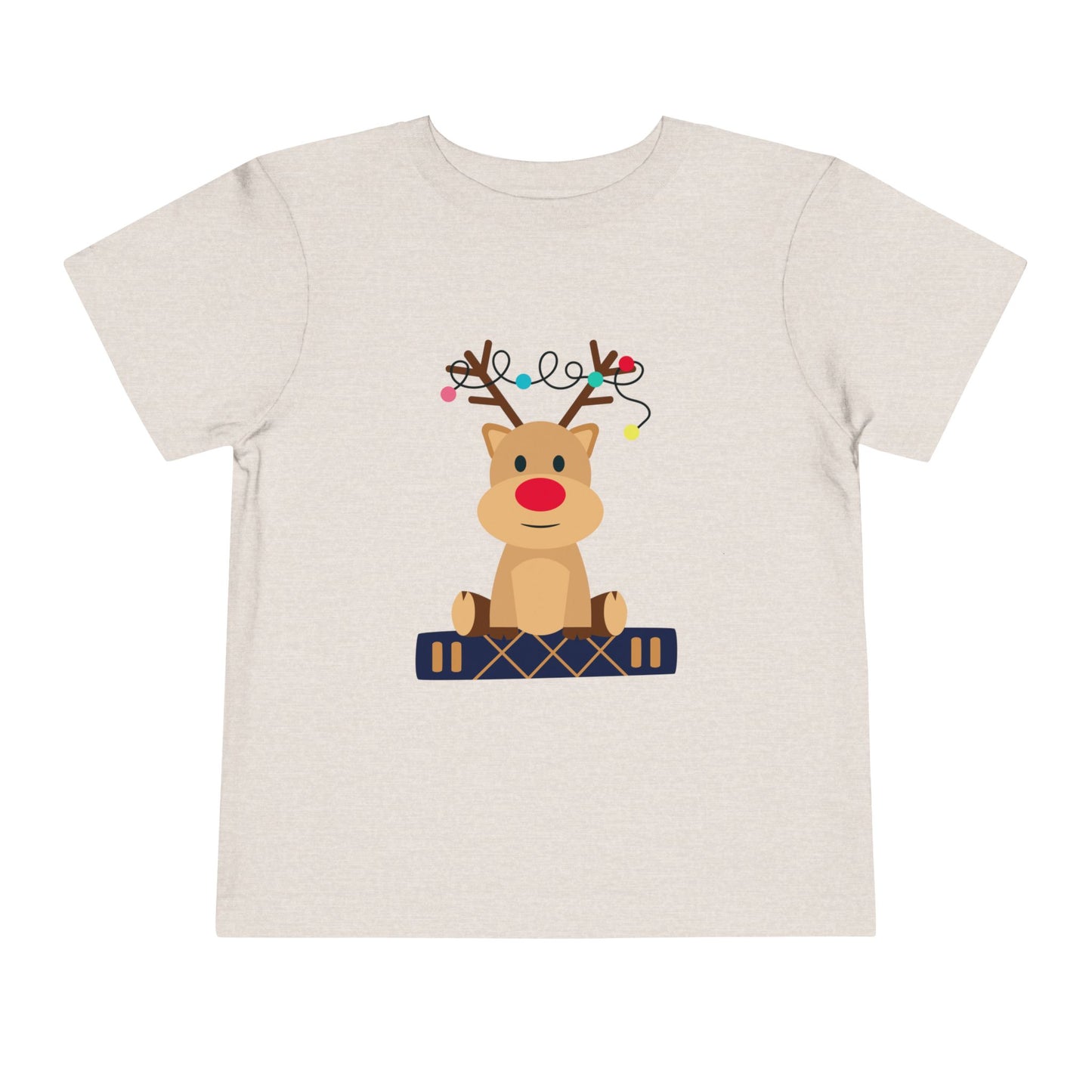 Beige/tan toddler t-shirt with a baby reindeer sitting on a book, a string of Christmas lights in it's antlers, on a white background