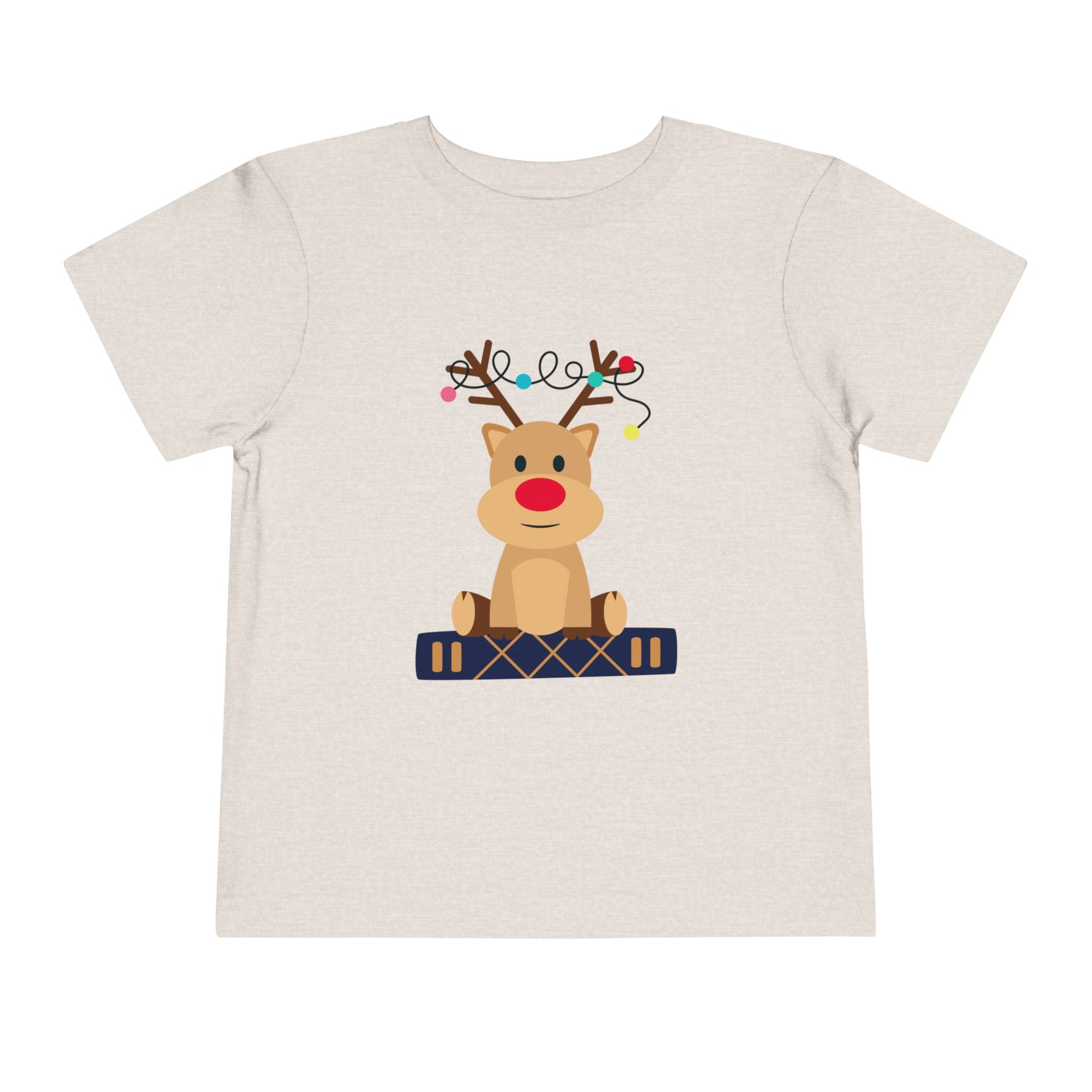 Beige/tan toddler t-shirt with a baby reindeer sitting on a book, a string of Christmas lights in it's antlers, on a white background