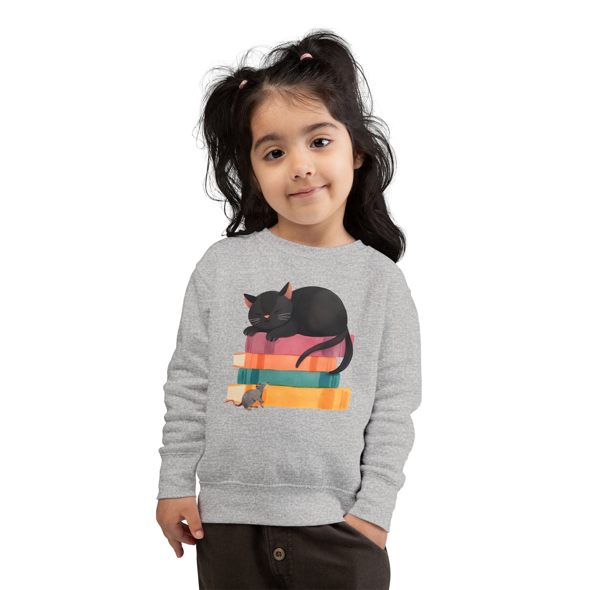 Girl wearing a gray toddler sweatshirt with a mouse looking up at a black cat sleeping on top of a stack of books 