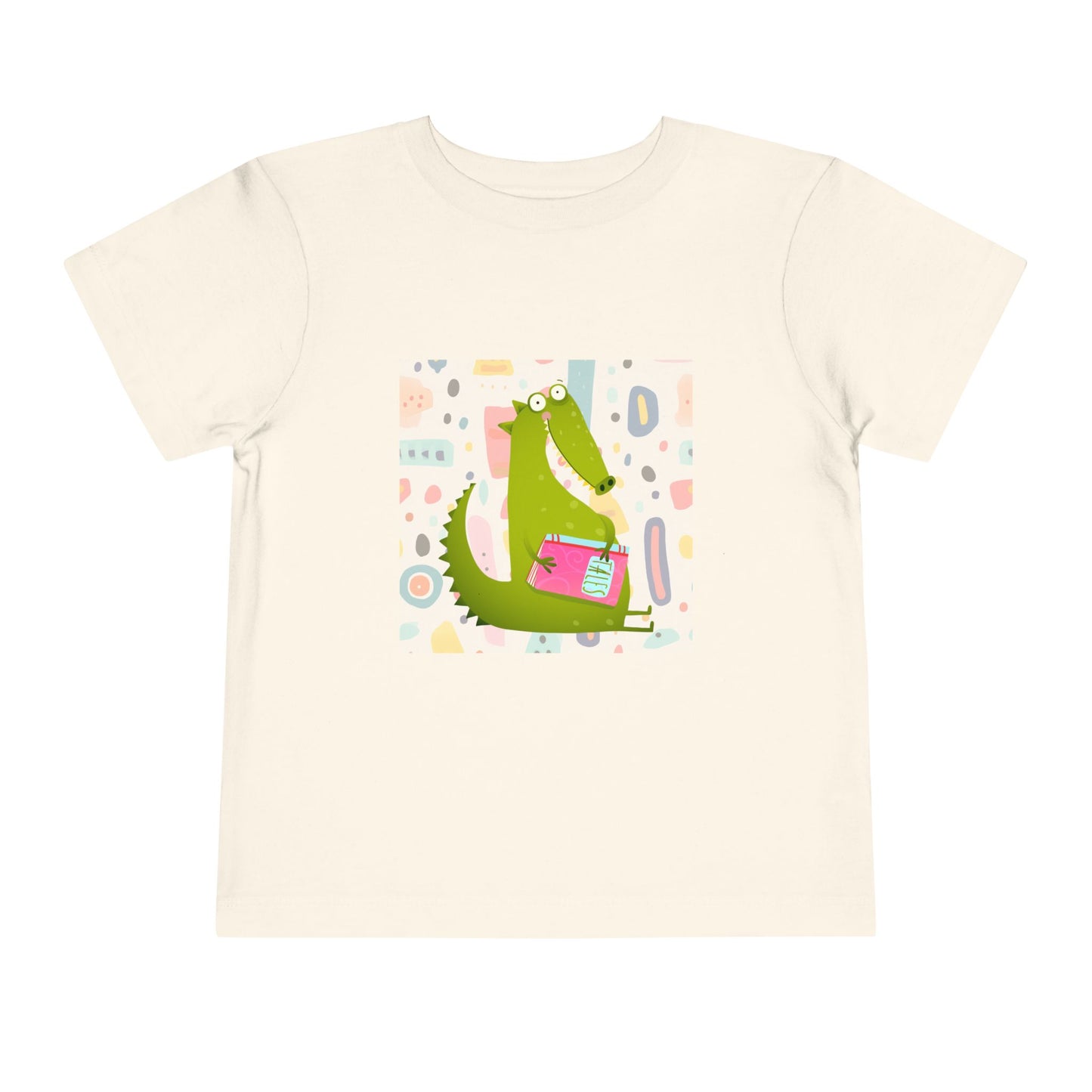 Beige toddler t-shirt with a green gator, holding pink book and colorful pastel background 