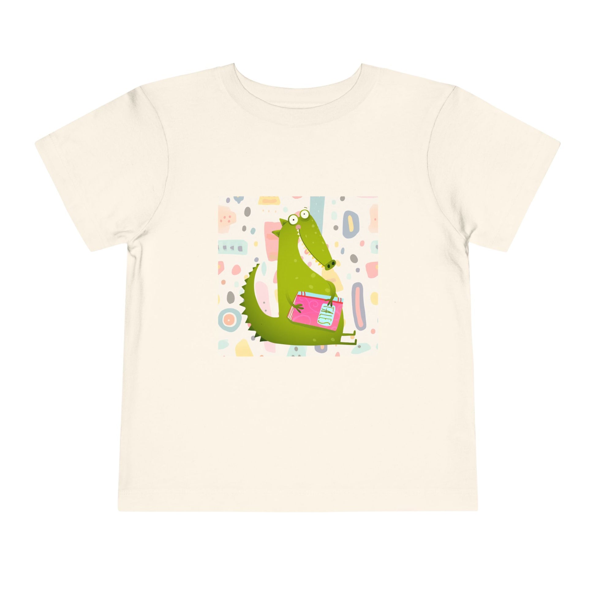 Beige toddler t-shirt with a green gator, holding pink book and colorful pastel background 