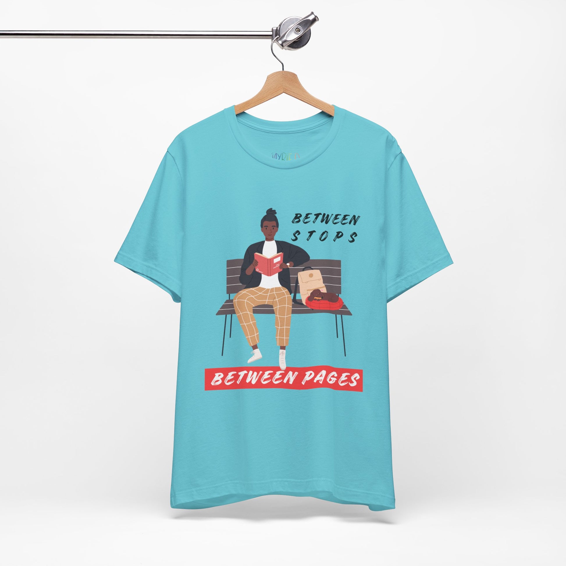 Turquoise t-shirt with a graphic of a person sitting on a bench reading a book, with text 'Between Stops Between Pages' 