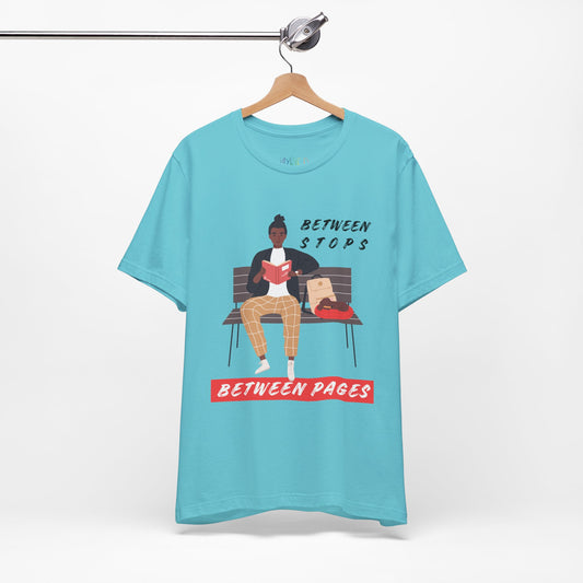 Turquoise t-shirt with a graphic of a person sitting on a bench reading a book, with text 'Between Stops Between Pages' 