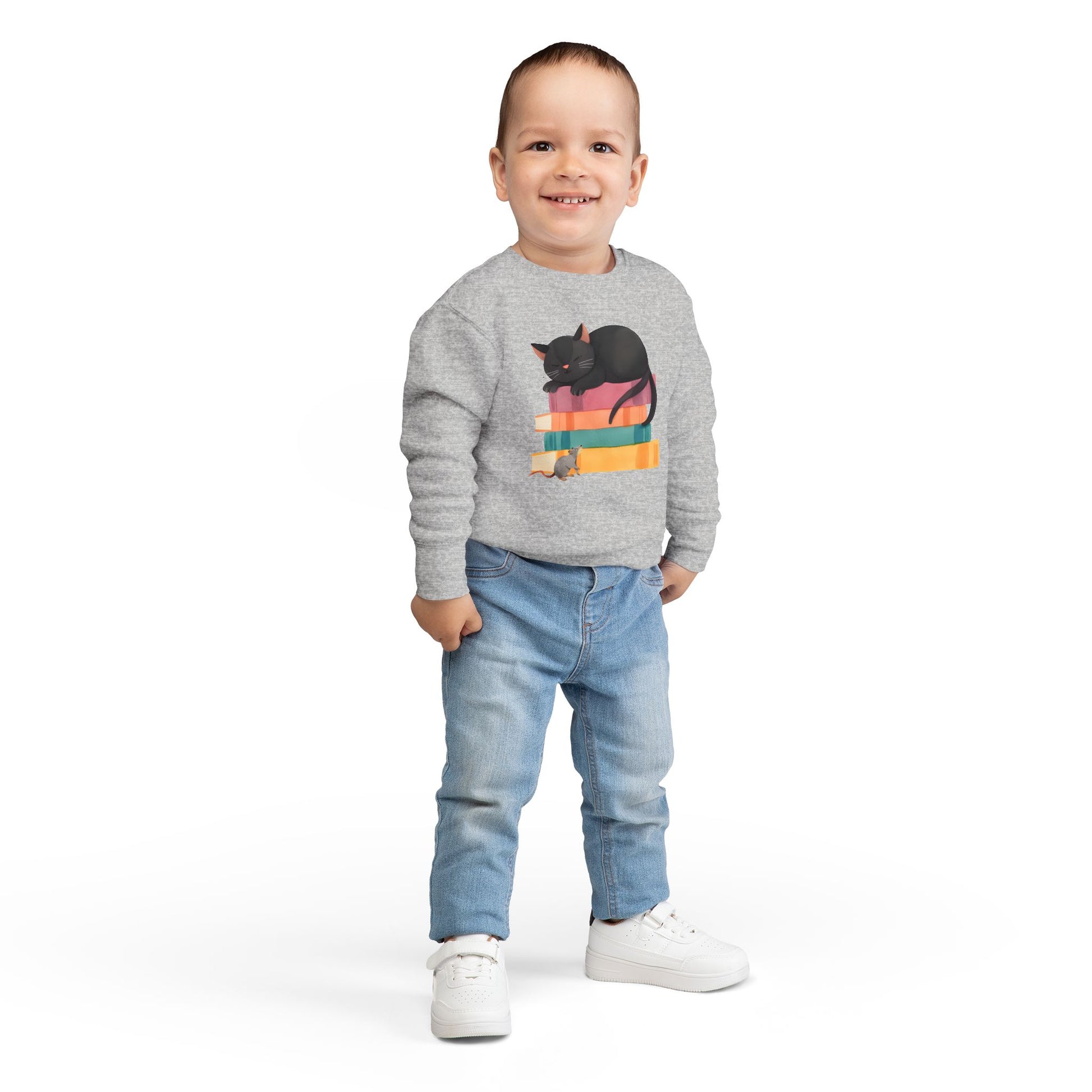 Boy wearing a gray toddler sweatshirt with a mouse looking up at a black cat sleeping on top of a stack of books 