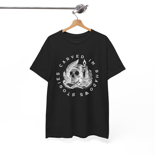 Black t-shirt with white graphic design of a creepy skull, books, candle and feathers with text 'Stories Carved In Shadows' on a hanger against a white background