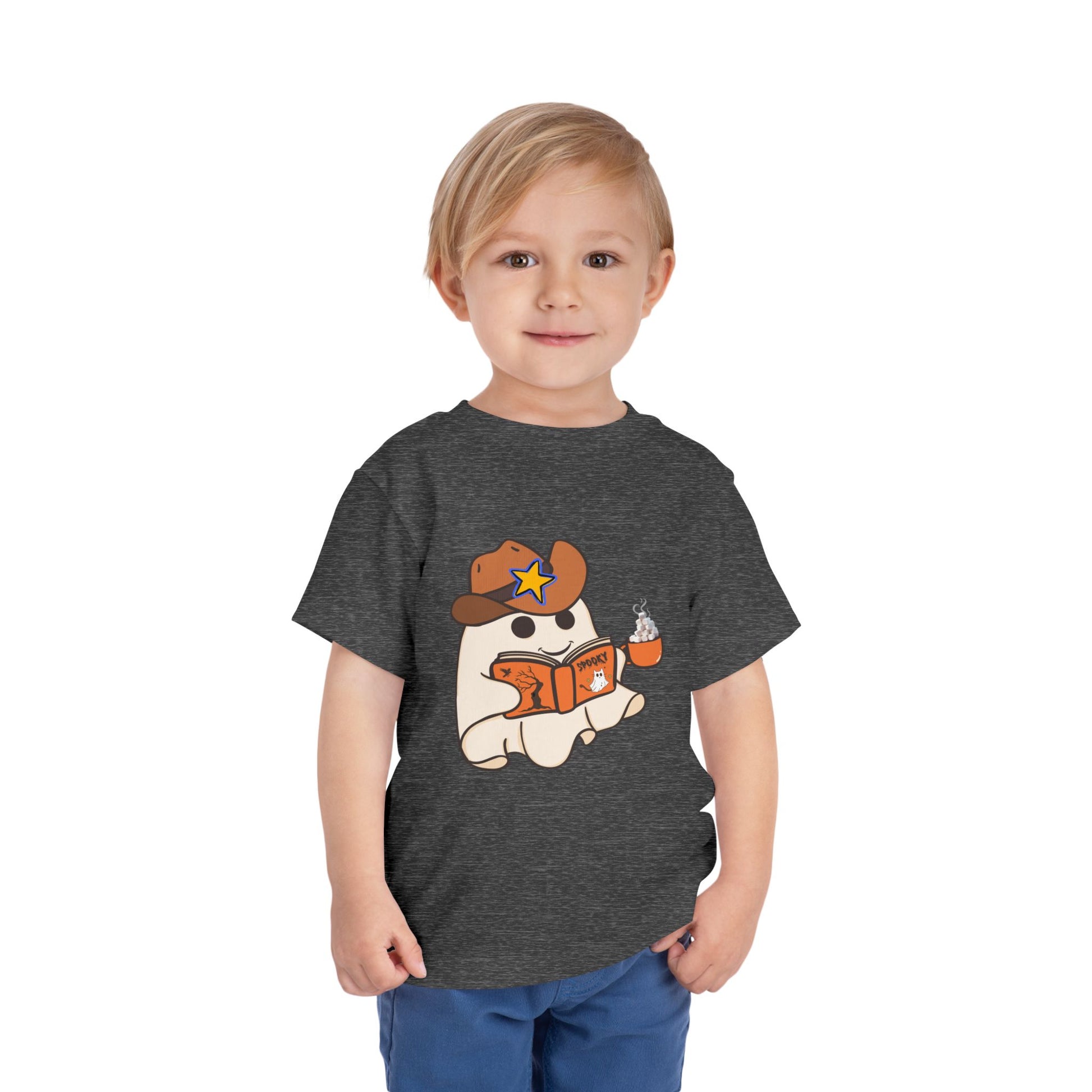 Child wearing a gray toddler t-shirt with a cartoon ghost, wearing a cowboy hat, reading a spooky book and holding a cup of hot cocoa
