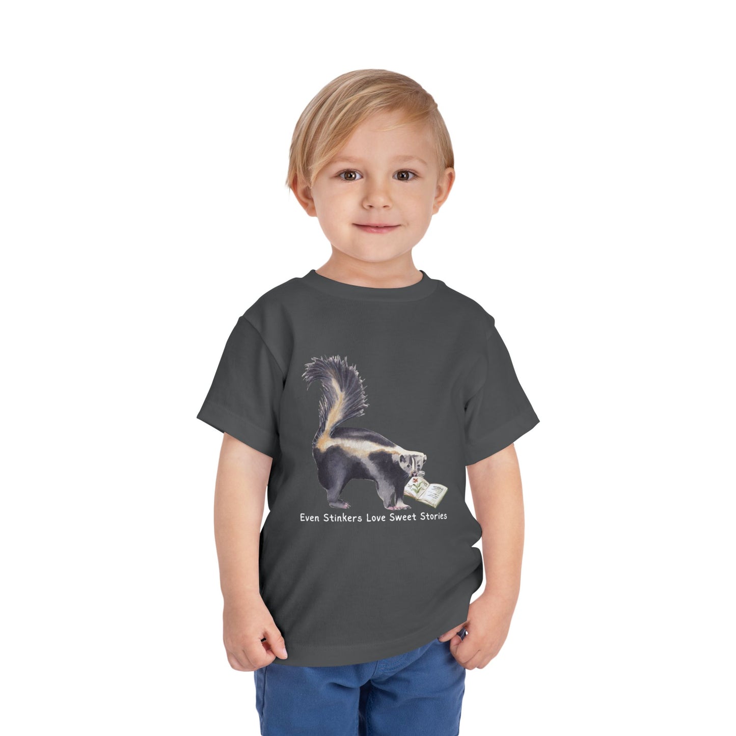 Child wearing a gray toddler t-shirt with a skunk reading a book and text 'Even Stinkers Love Sweet Stories' 