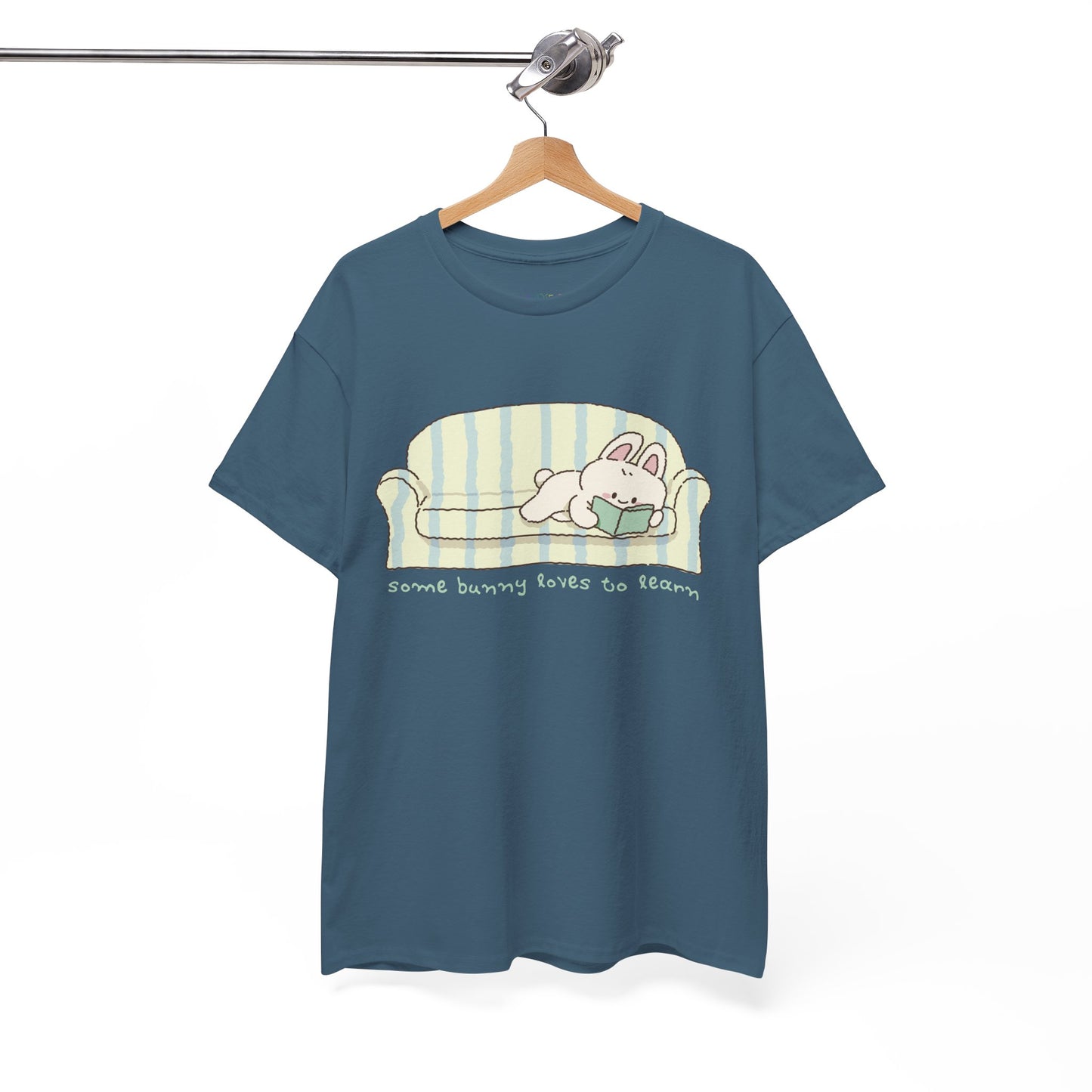 Blue t-shirt with a cartoon bunny reading a book as it's lays on a couch, text 'some bunny loves to learn' 