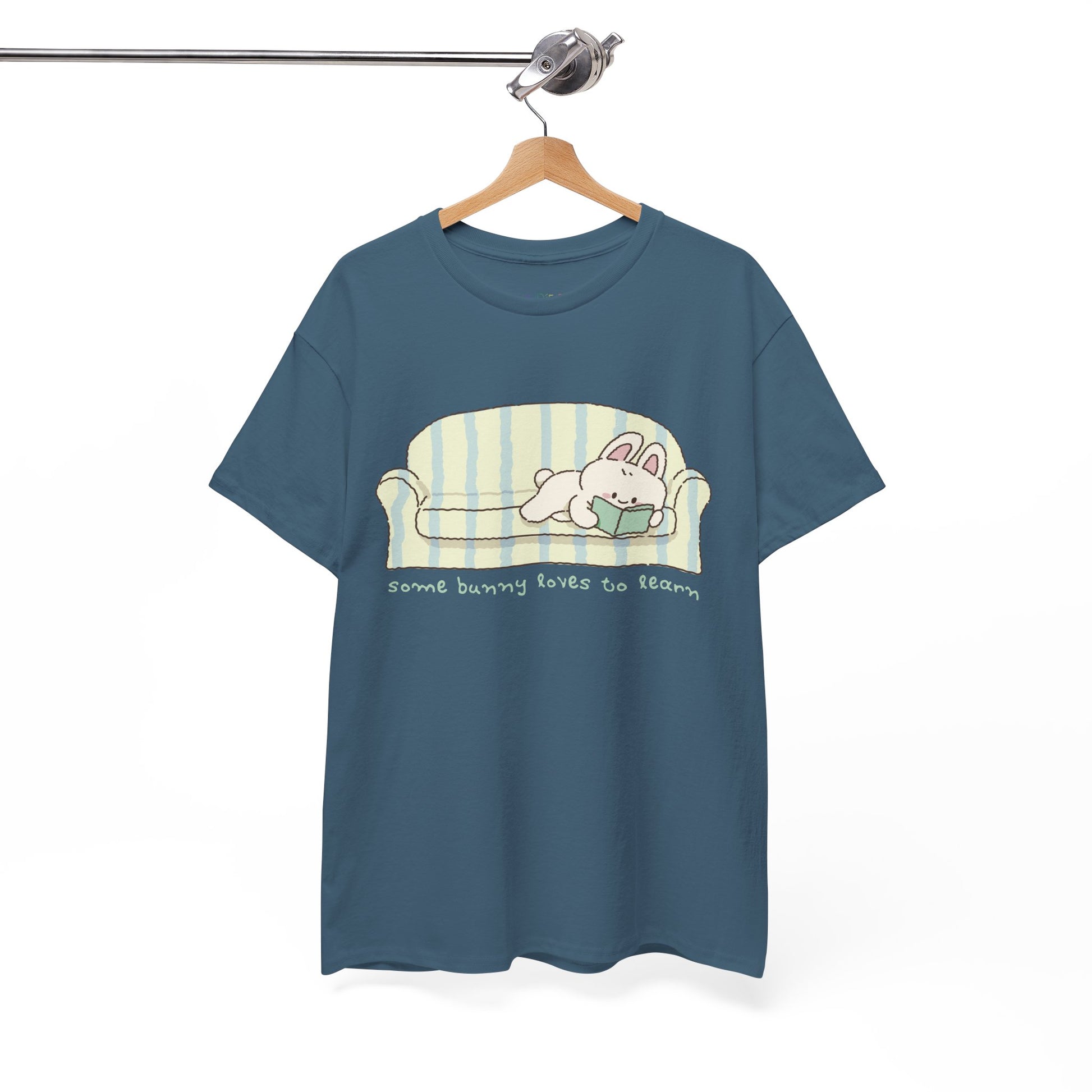 Blue t-shirt with a cartoon bunny reading a book as it's lays on a couch, text 'some bunny loves to learn' 