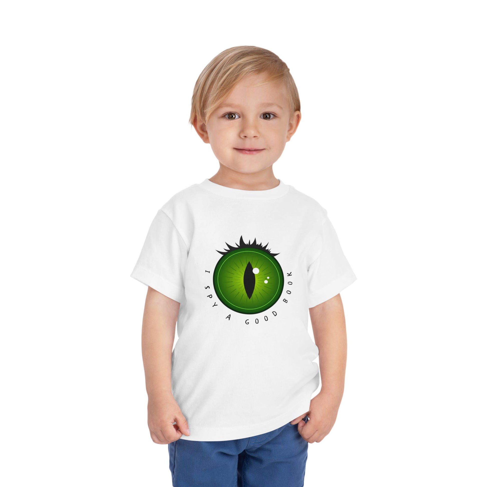 Toddler wearing a white t-shirt with a green dragon eye design and text 'I SPY A GOOD BOOK'