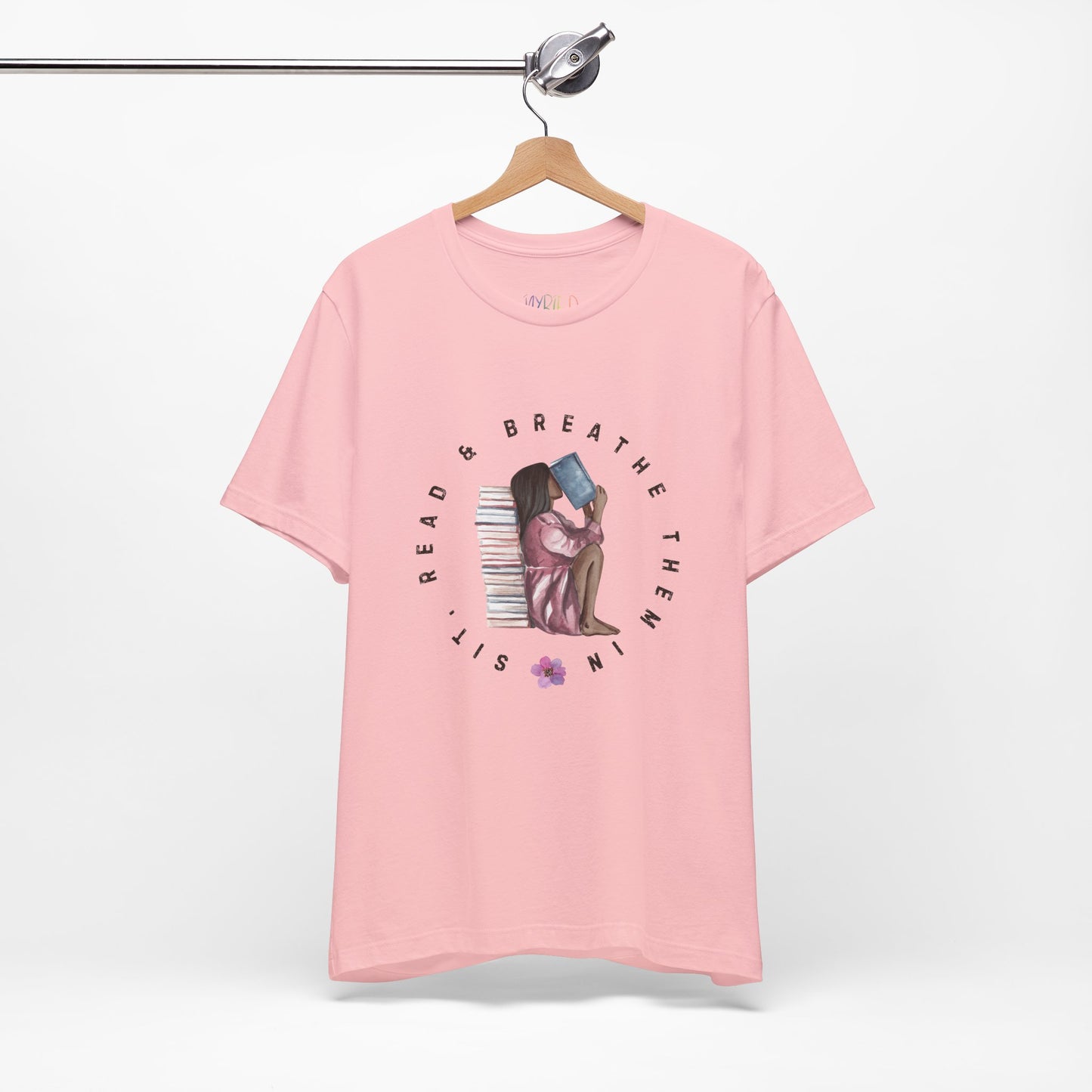 Pink t-shirt with graphic design of a girl sitting on the ground against a stack of books, inhaling the book she's reading and text 'Sit, Read & Breathe Them In'