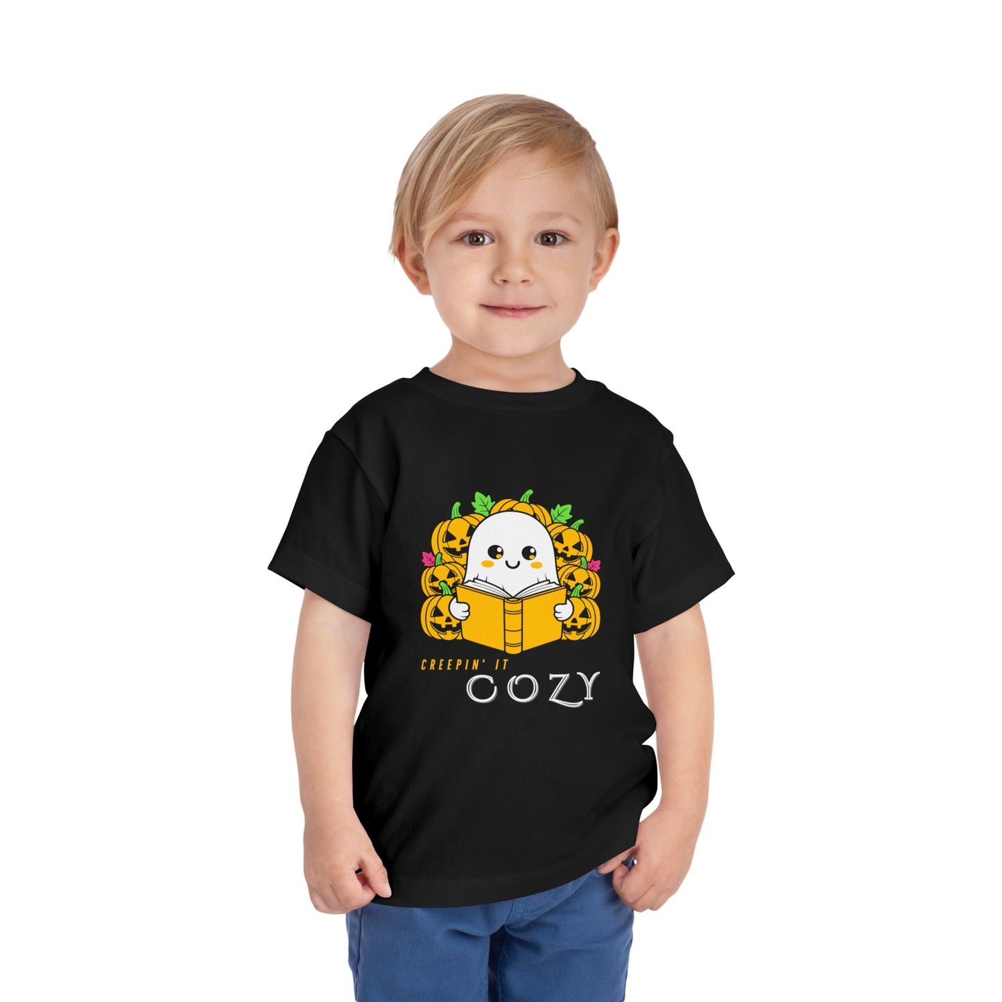 Child wearing a black toddler t-shirt with a ghost reading a book in pumpkin patch and text 'Creepin' It Cozy'