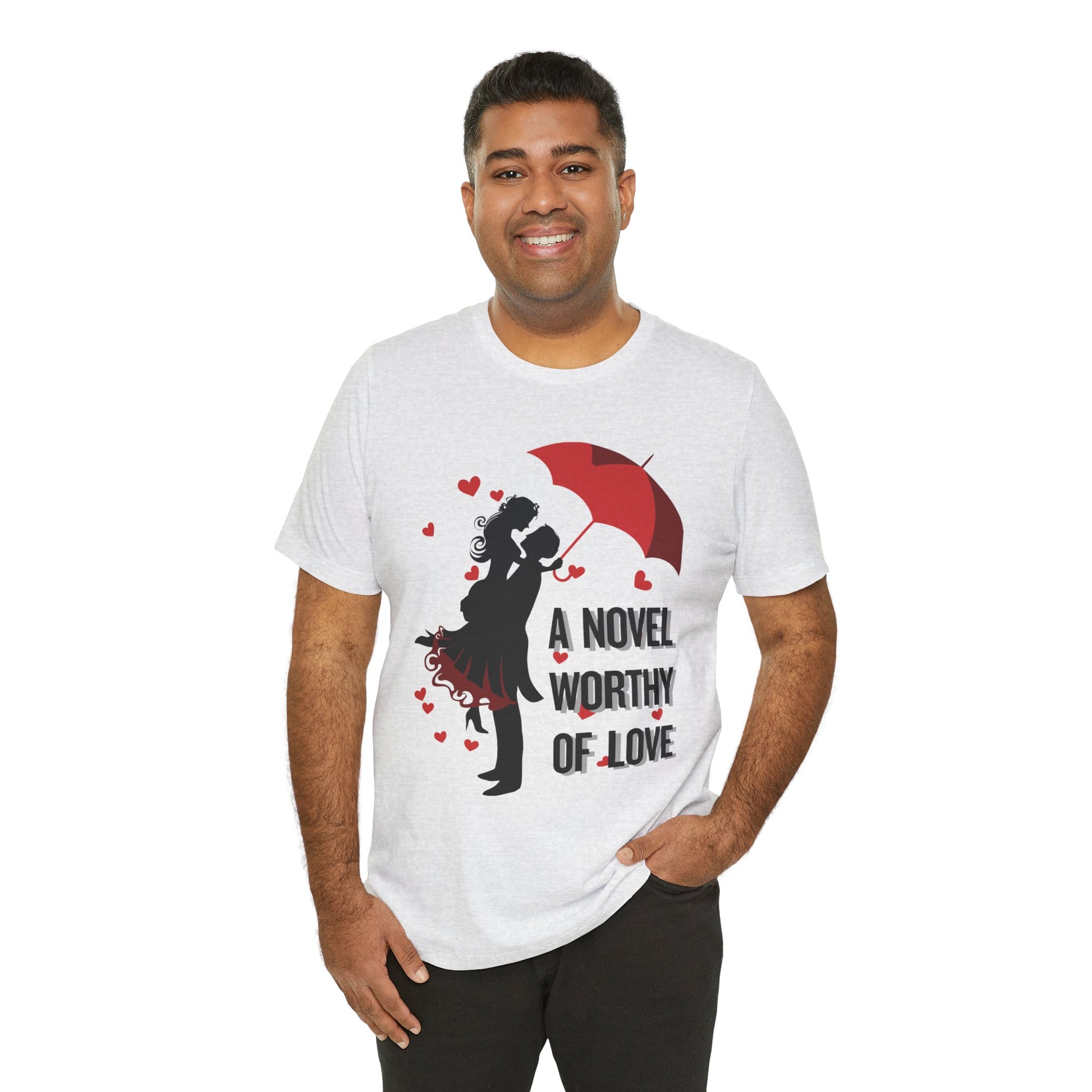 A man wearing an ash t-shirt with a black graphic design of a man holding a woman under a red umbrella, sprinkled with hearts and text 'A Novel Worthy of Love' 