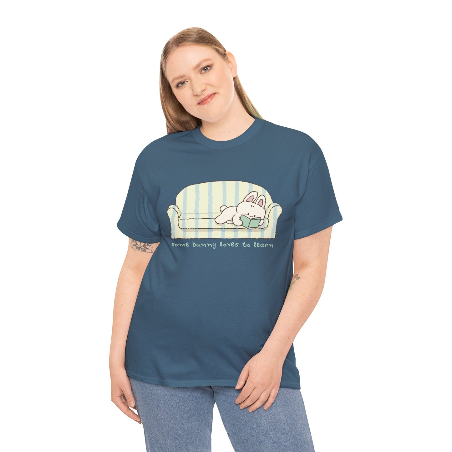 Woman wearing a blue t-shirt with a cartoon bunny reading a book as it's lays on a couch, text 'some bunny loves to learn' 