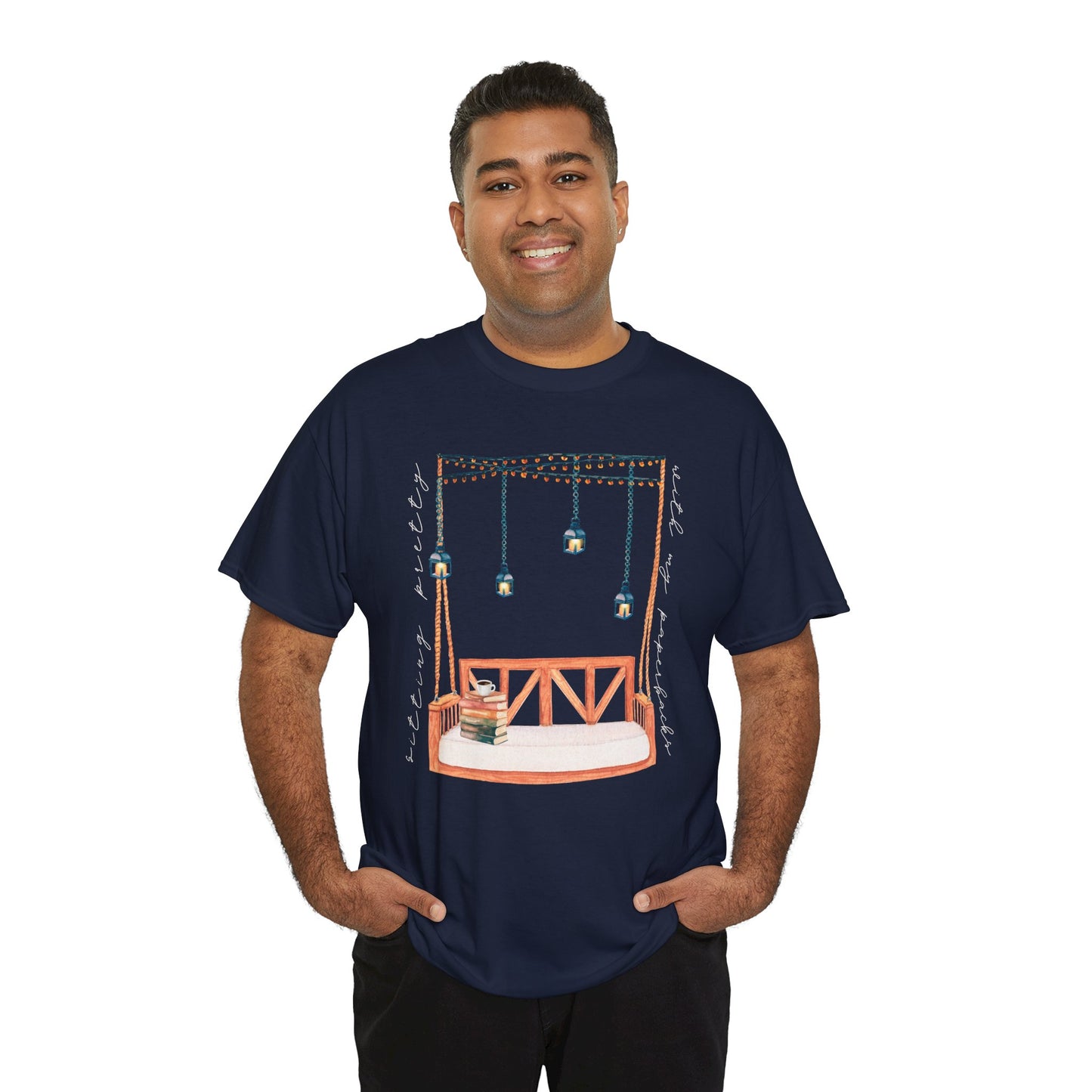 Man wearing a navy blue t-shirt with a porch swing, string lights, hot beverage and a stack of books, text says 'Sitting Pretty With My Paperbacks'
