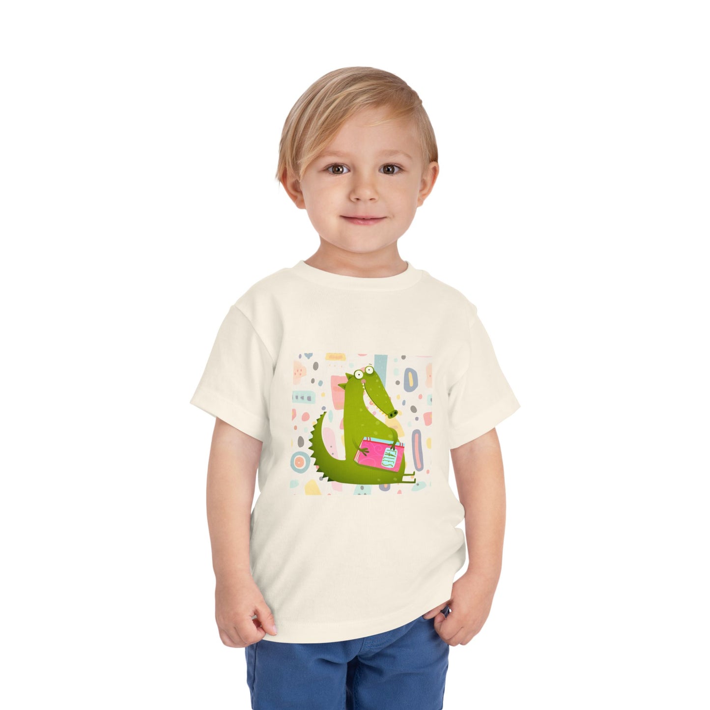 Child wearing a beige toddler t-shirt with a green gator, holding pink book and colorful pastel background 