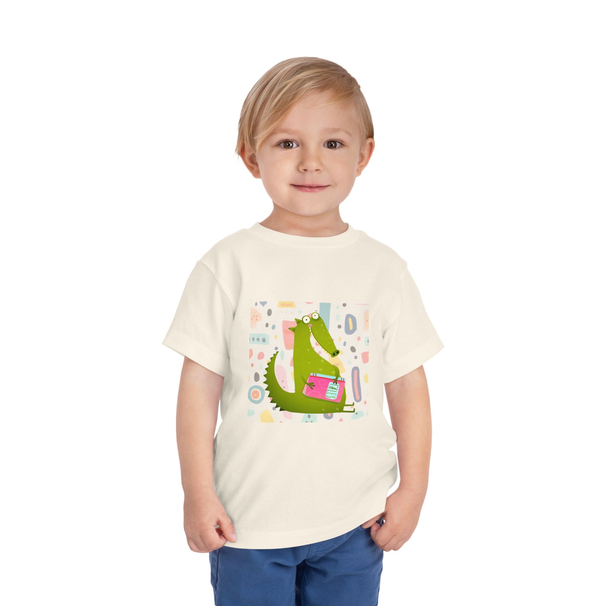 Child wearing a beige toddler t-shirt with a green gator, holding pink book and colorful pastel background 