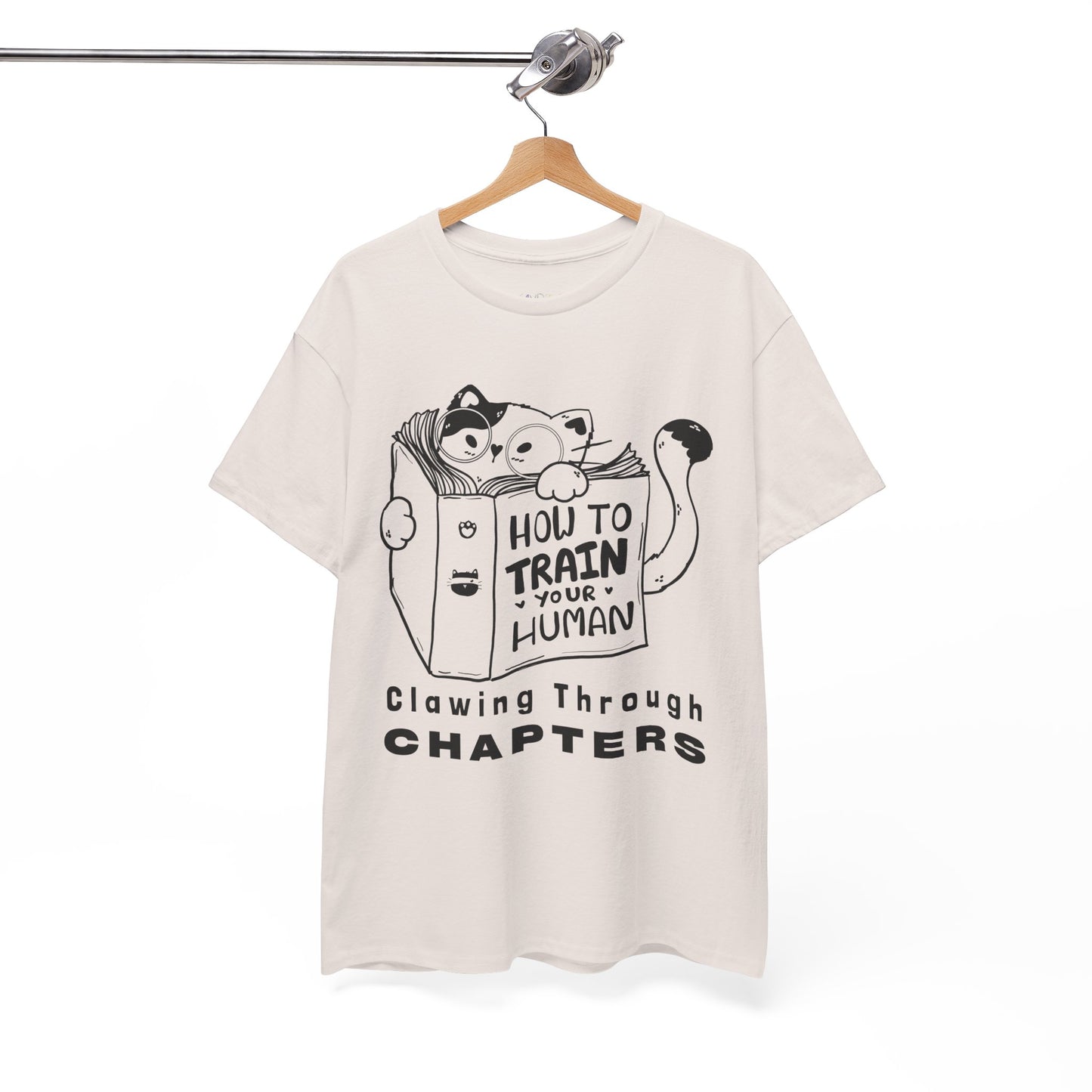 Beige t-shirt with black graphic design of a cat reading a 'How To Train Your Human' book and text 'Clawing Through Chapters'