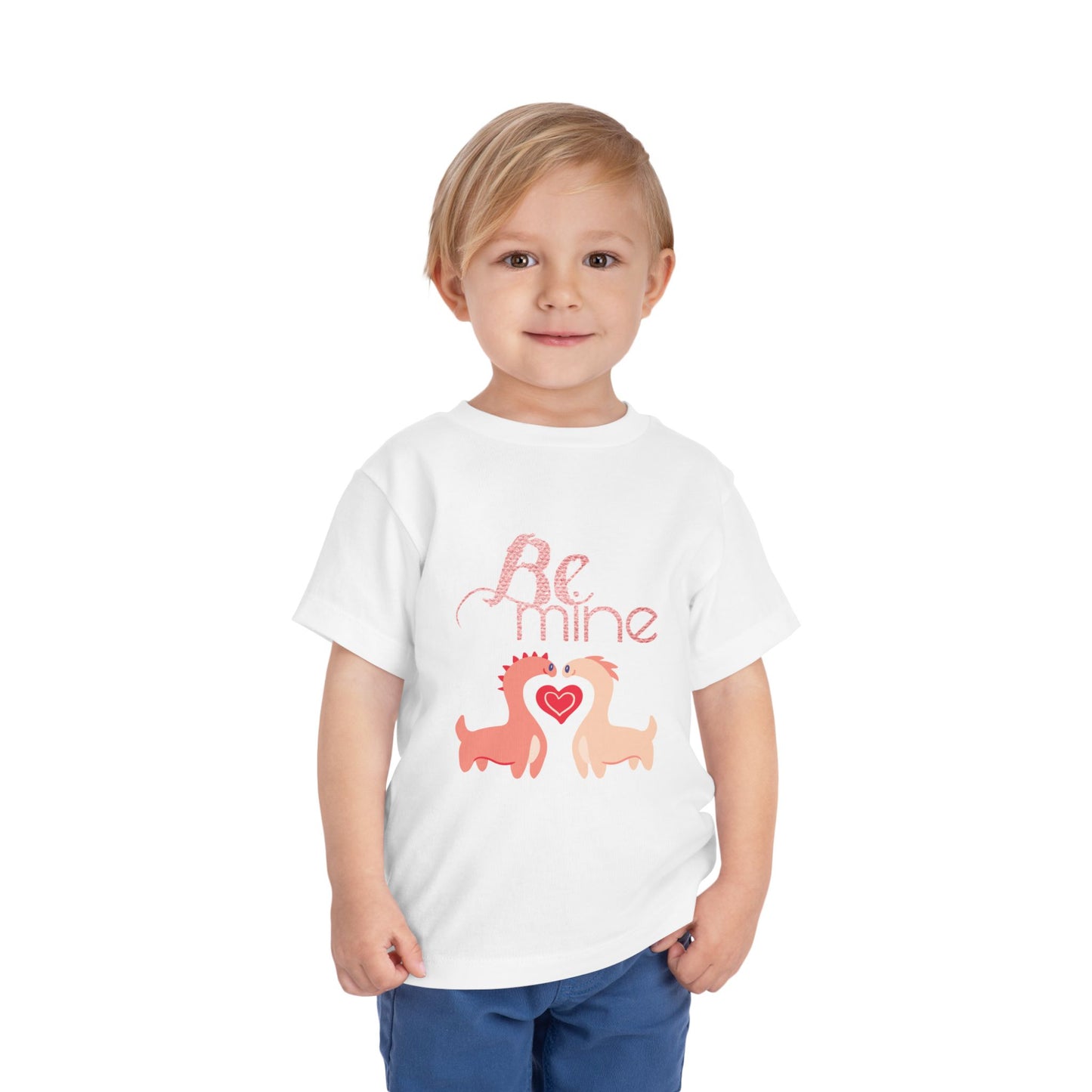 Child wearing a white toddler t-shirt with two pink dinosaurs, a heart and 'Be Mine' text 