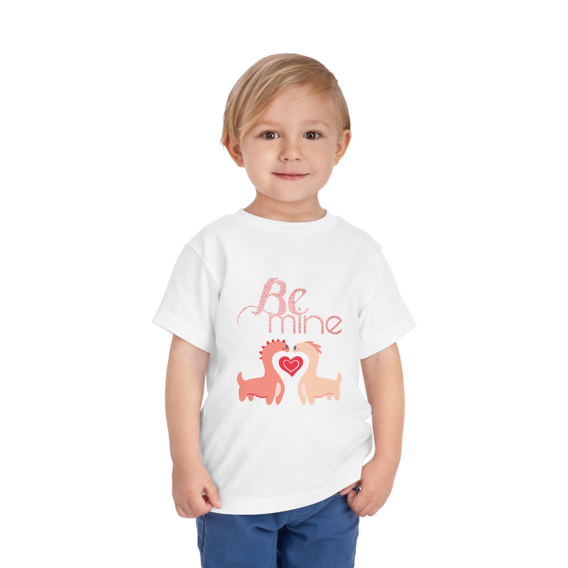 Child wearing a white toddler t-shirt with two pink dinosaurs, a heart and 'Be Mine' text 