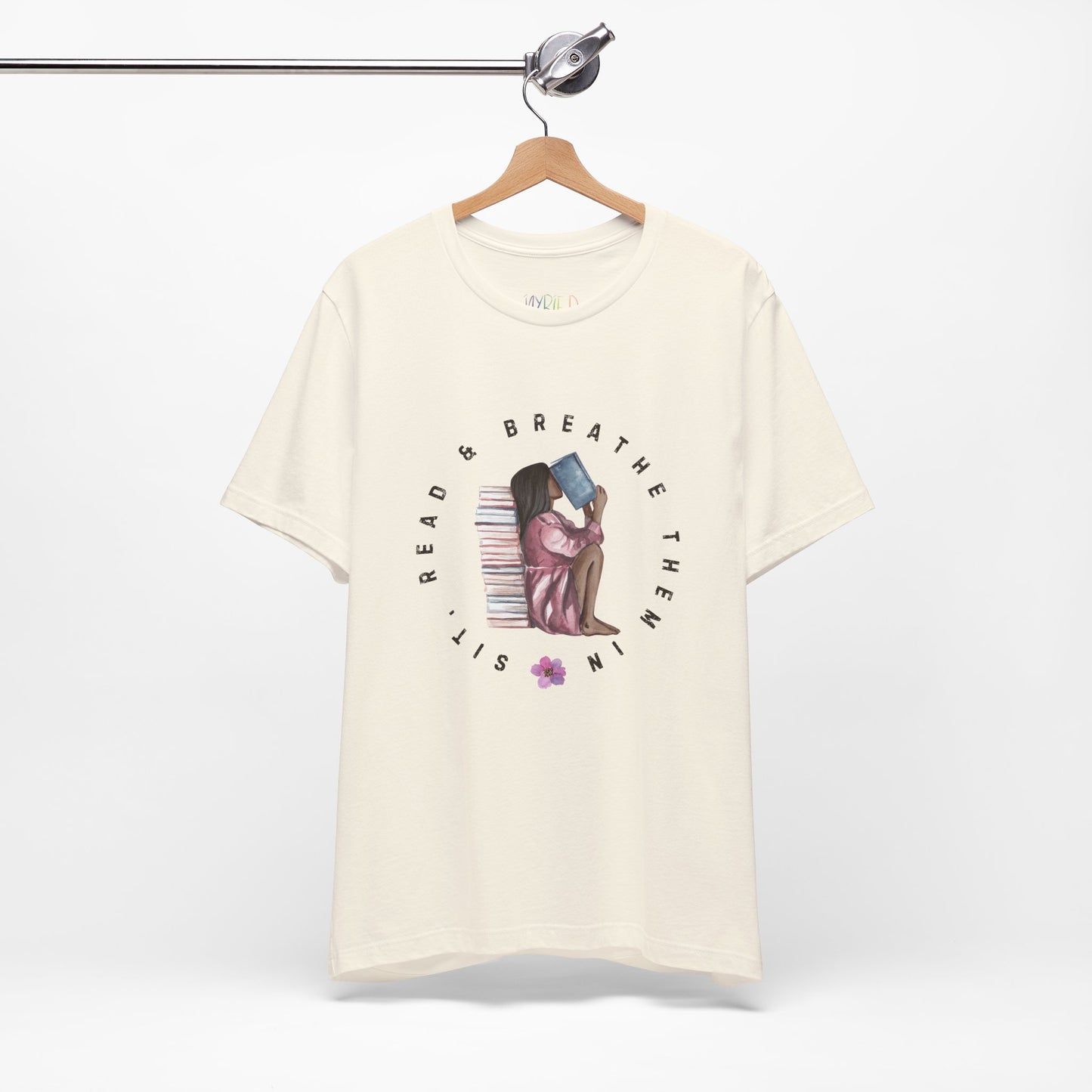Beige t-shirt with graphic design of a girl sitting on the ground against a stack of books, inhaling the book she's reading and text 'Sit, Read & Breathe Them In'