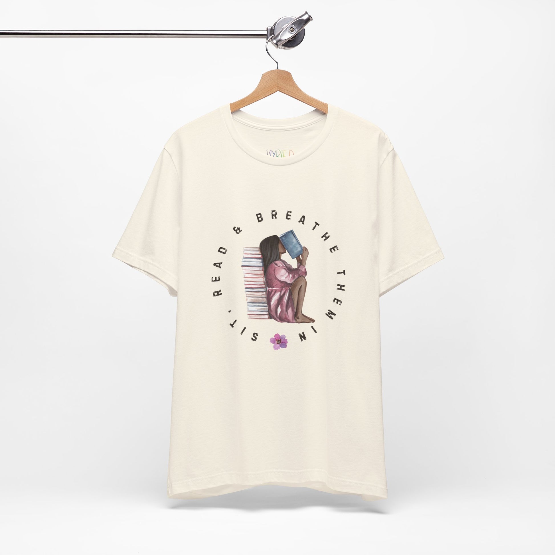 Beige t-shirt with graphic design of a girl sitting on the ground against a stack of books, inhaling the book she's reading and text 'Sit, Read & Breathe Them In'