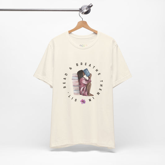 Beige t-shirt with graphic design of a girl sitting on the ground against a stack of books, inhaling the book she's reading and text 'Sit, Read & Breathe Them In'