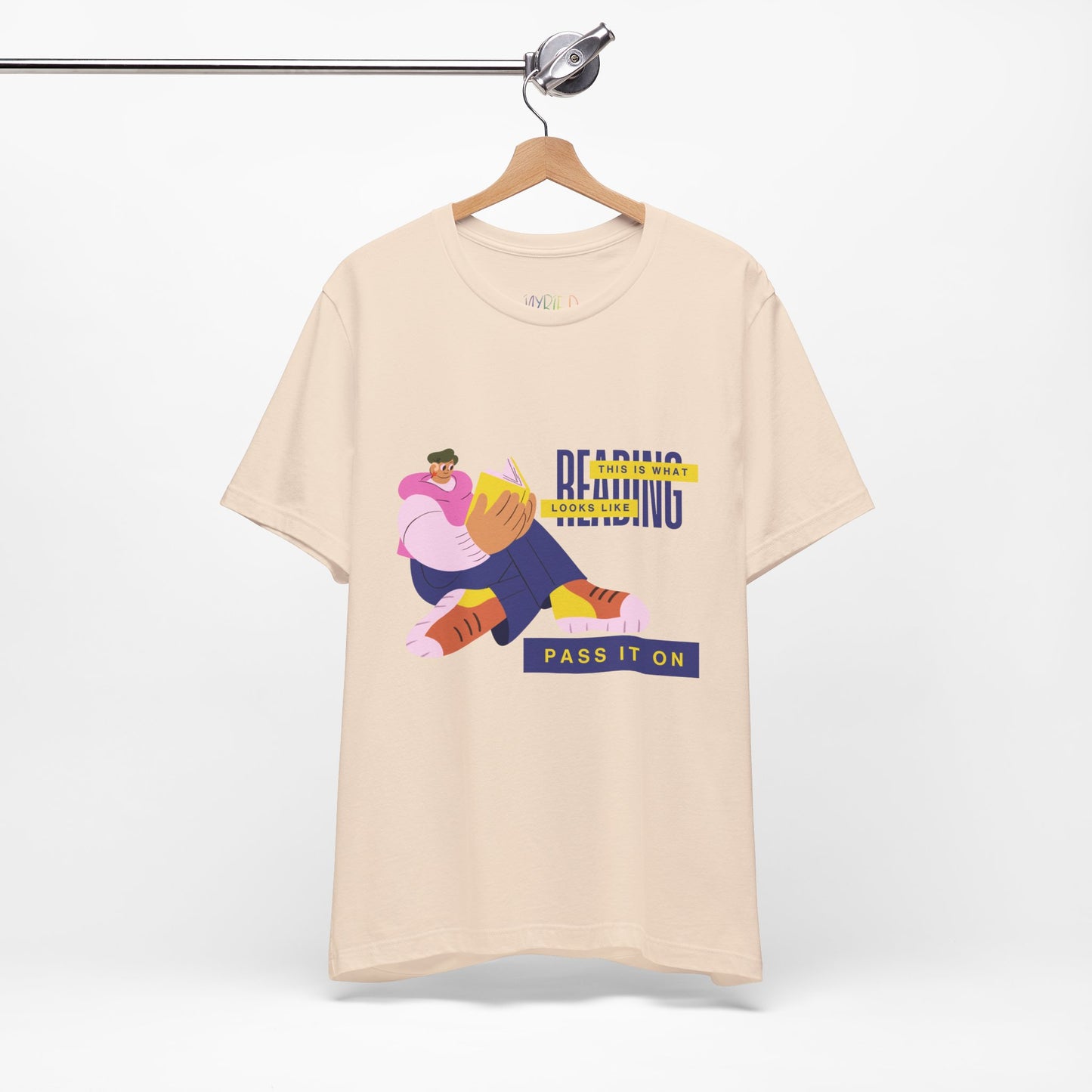 Cream t-shirt with colorful, funny shaped figure reading a book and text 'This Is What Reading Looks Like, Pass It On'