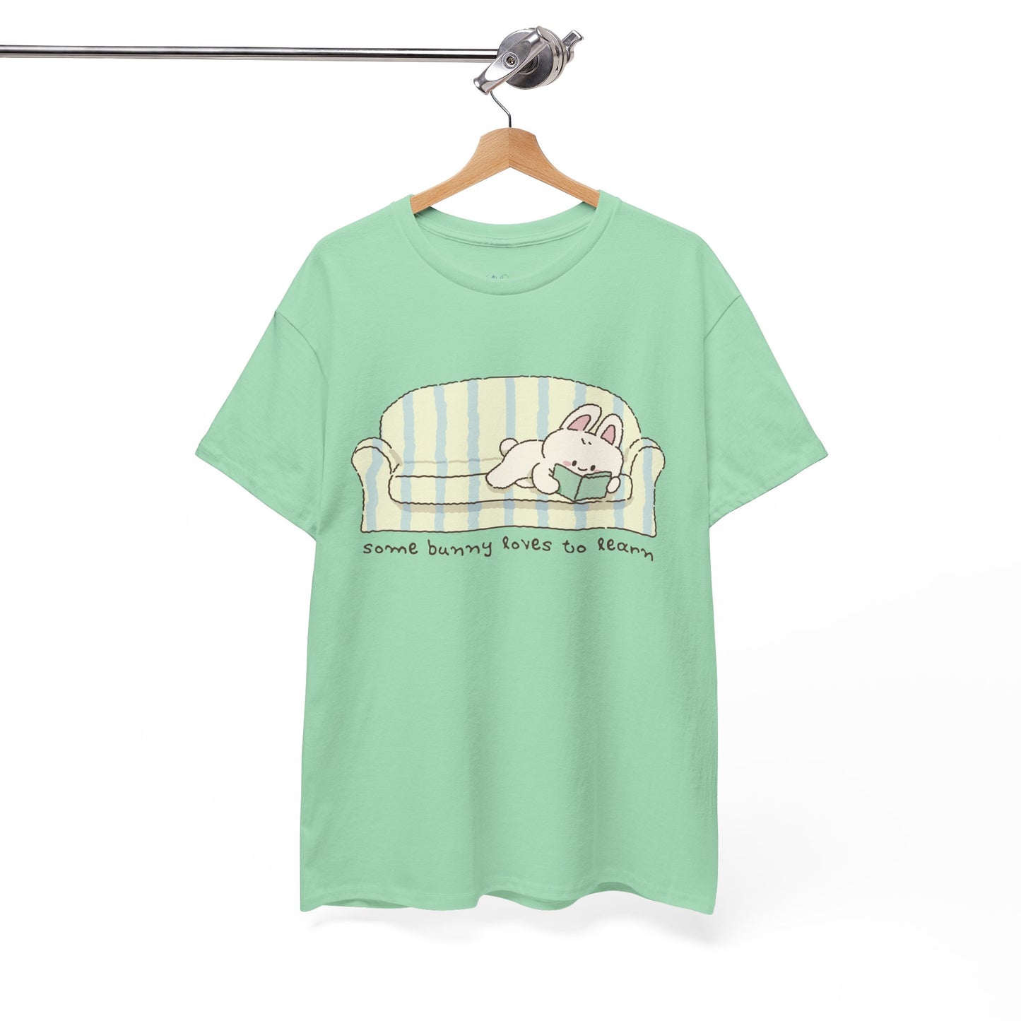 Light green t-shirt with a cartoon bunny reading a book as it's lays on a couch, text 'some bunny loves to learn' 