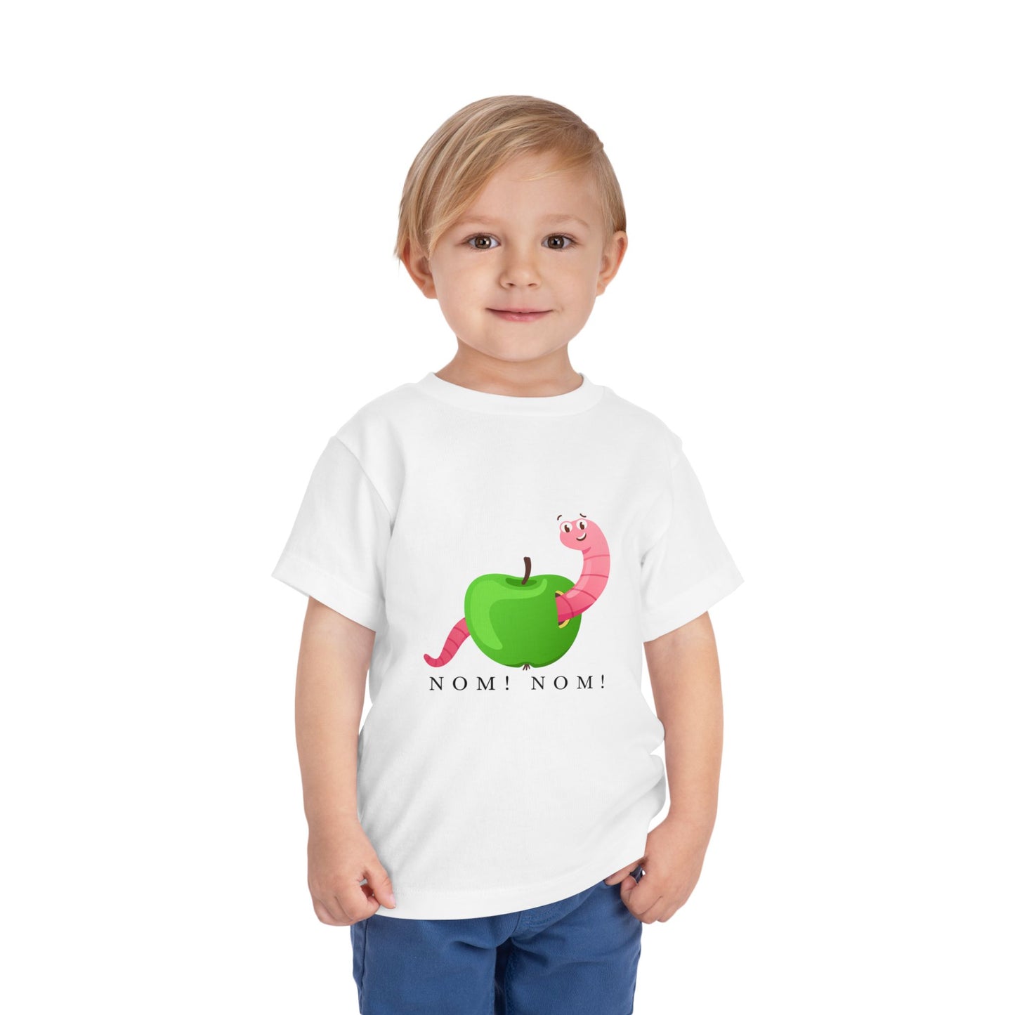 Child wearing a white toddler t-shirt with a green apple and pink worm graphic and 'NOM! NOM!' text 