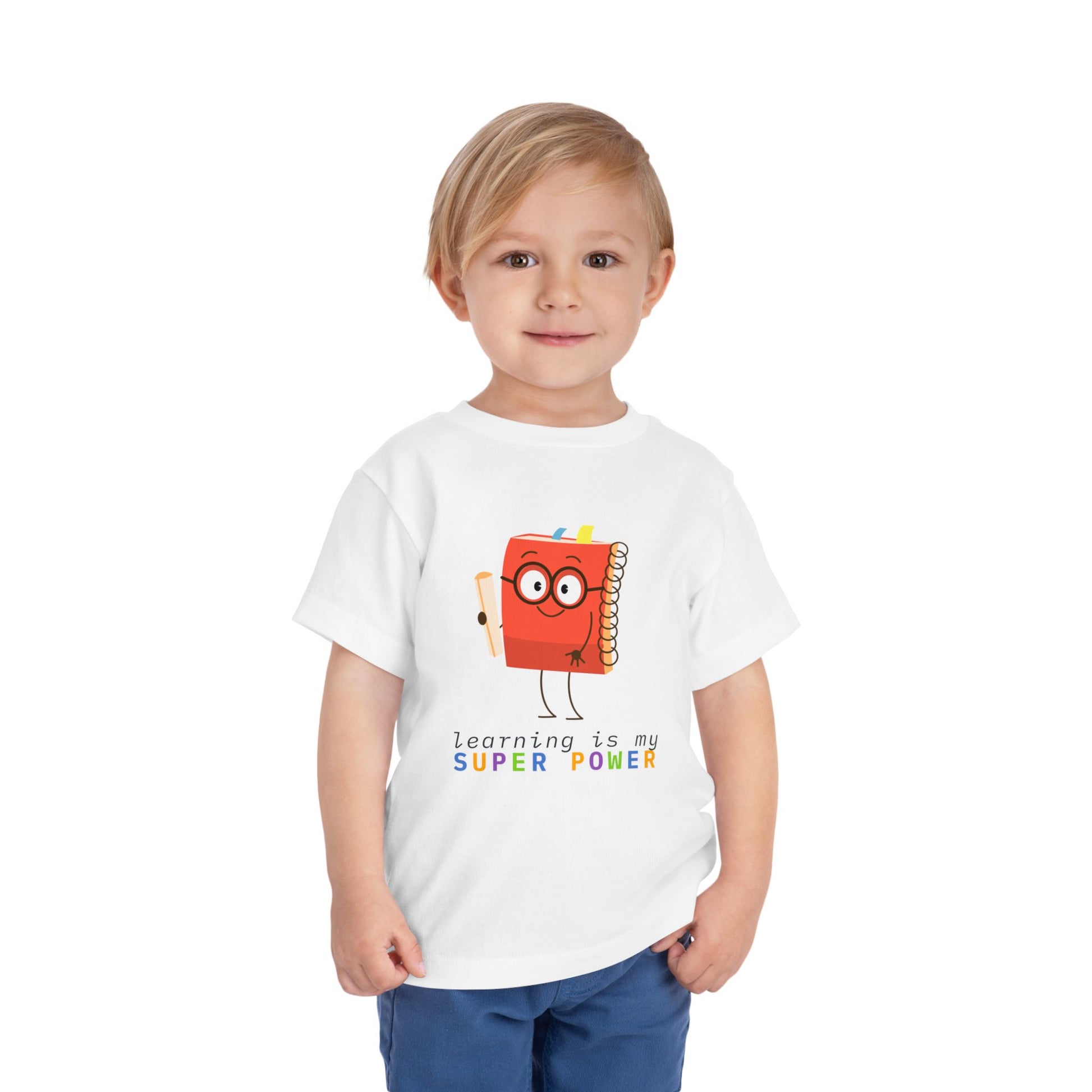 Child wearing a white toddler t-shirt with a cartoon book character and colorful 'learning is my super power' text 
