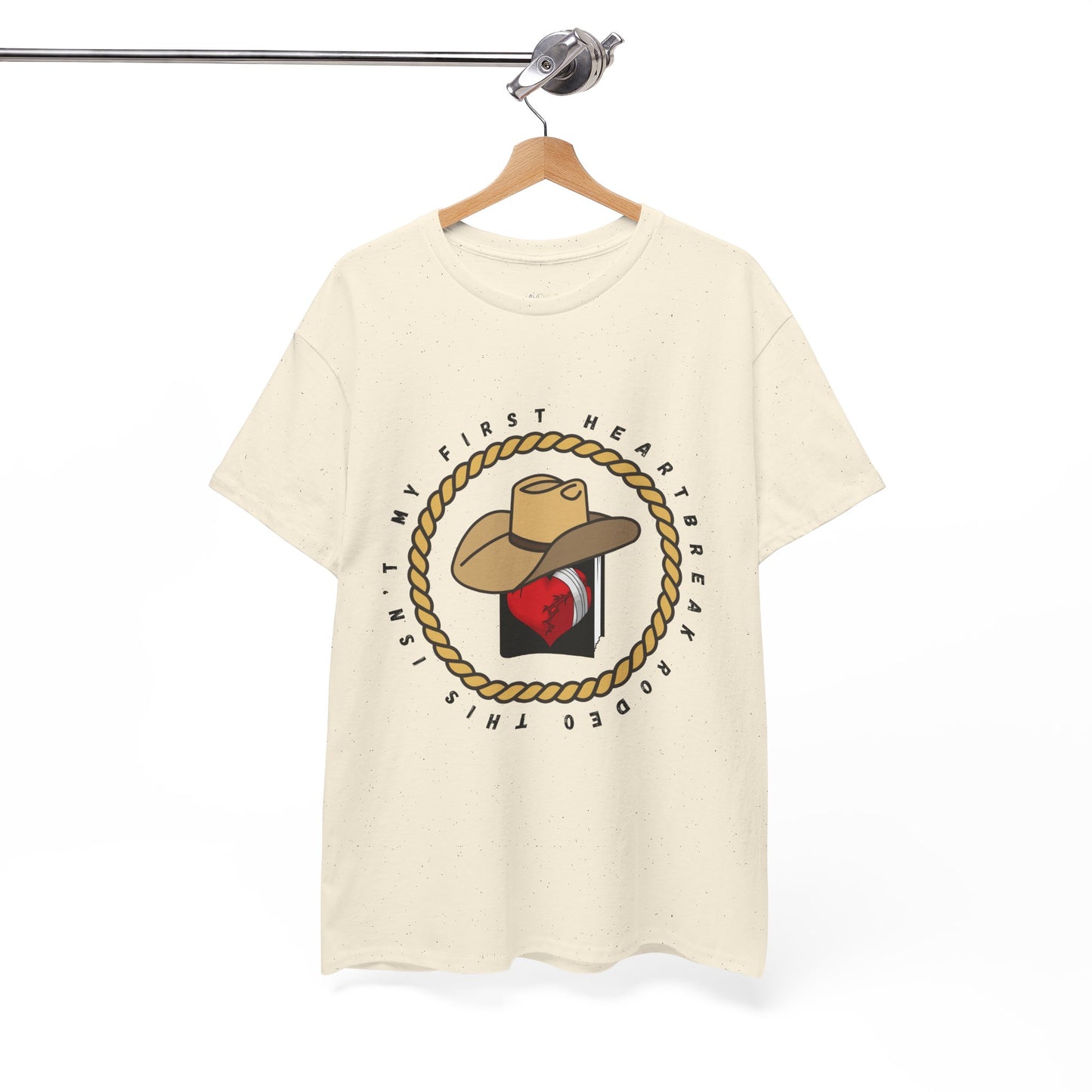 Beige t-shirt with a cowboy hat atop a book with a broken heart, circled by a rope and text 'This Isn't My First Heartbreak Rodeo'