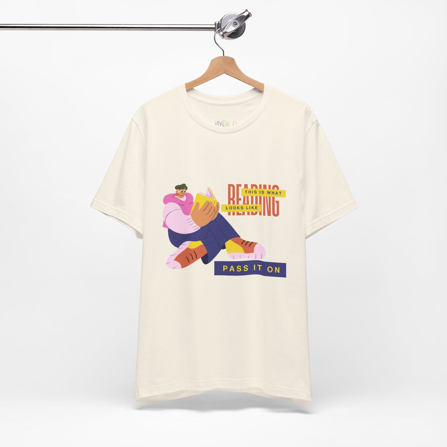 Beige t-shirt with colorful, funny shaped figure reading a book and text 'This Is What Reading Looks Like, Pass It On'