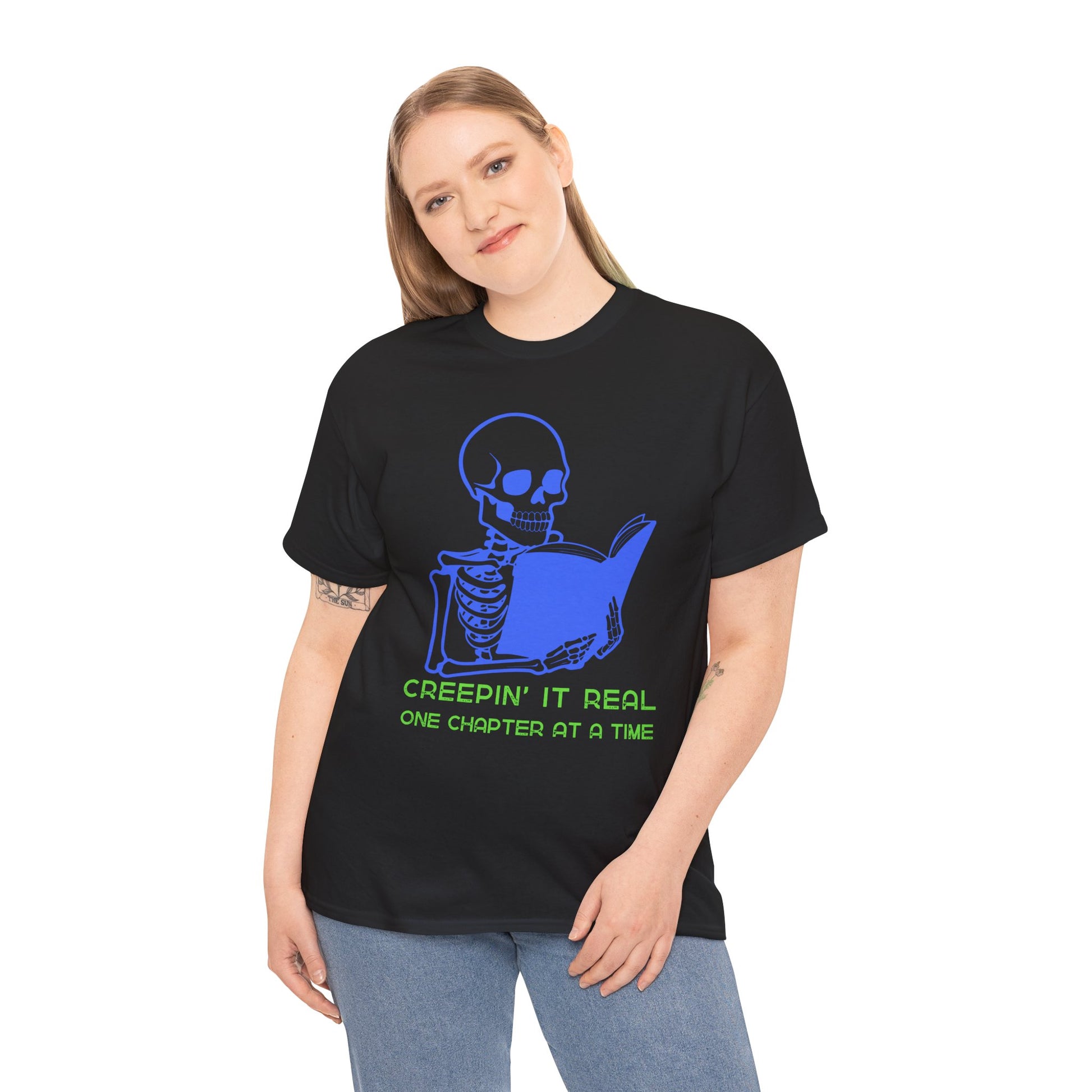 Woman wearing a black t-shirt with blue skeleton graphic reading a book and green text 'Creepin' It Real One Chapter At A Time' on a hanger against a white background