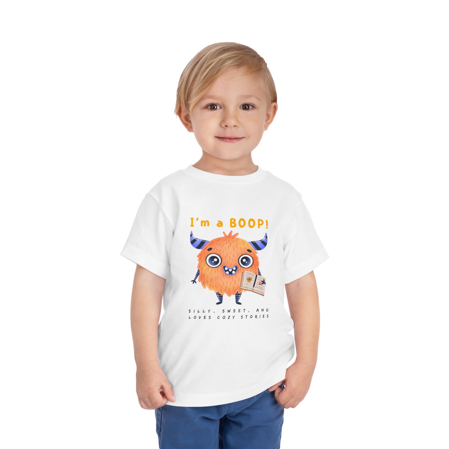 Child wearing a white toddler personality t-shirt with a cartoon monster holding a book, proudly proclaiming 'I'm A BOOP!' Sub-text "Silly, Sweet, And Loves Cozy Stories' on a white background