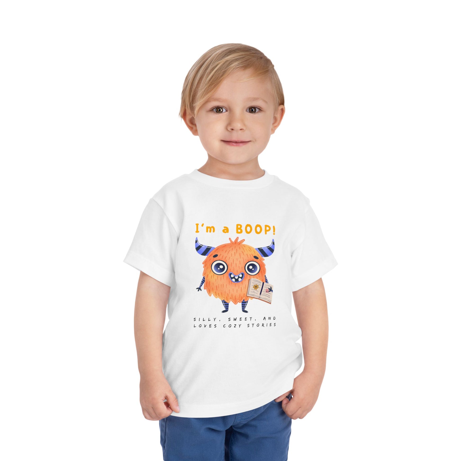 Child wearing a white toddler personality t-shirt with a cartoon monster holding a book, proudly proclaiming 'I'm A BOOP!' Sub-text "Silly, Sweet, And Loves Cozy Stories' on a white background
