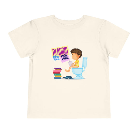 Beige t-shirt with a graphic of a child reading on a toilet, a stack of books at his feet, and colorful 'Reading Takes Time' text.